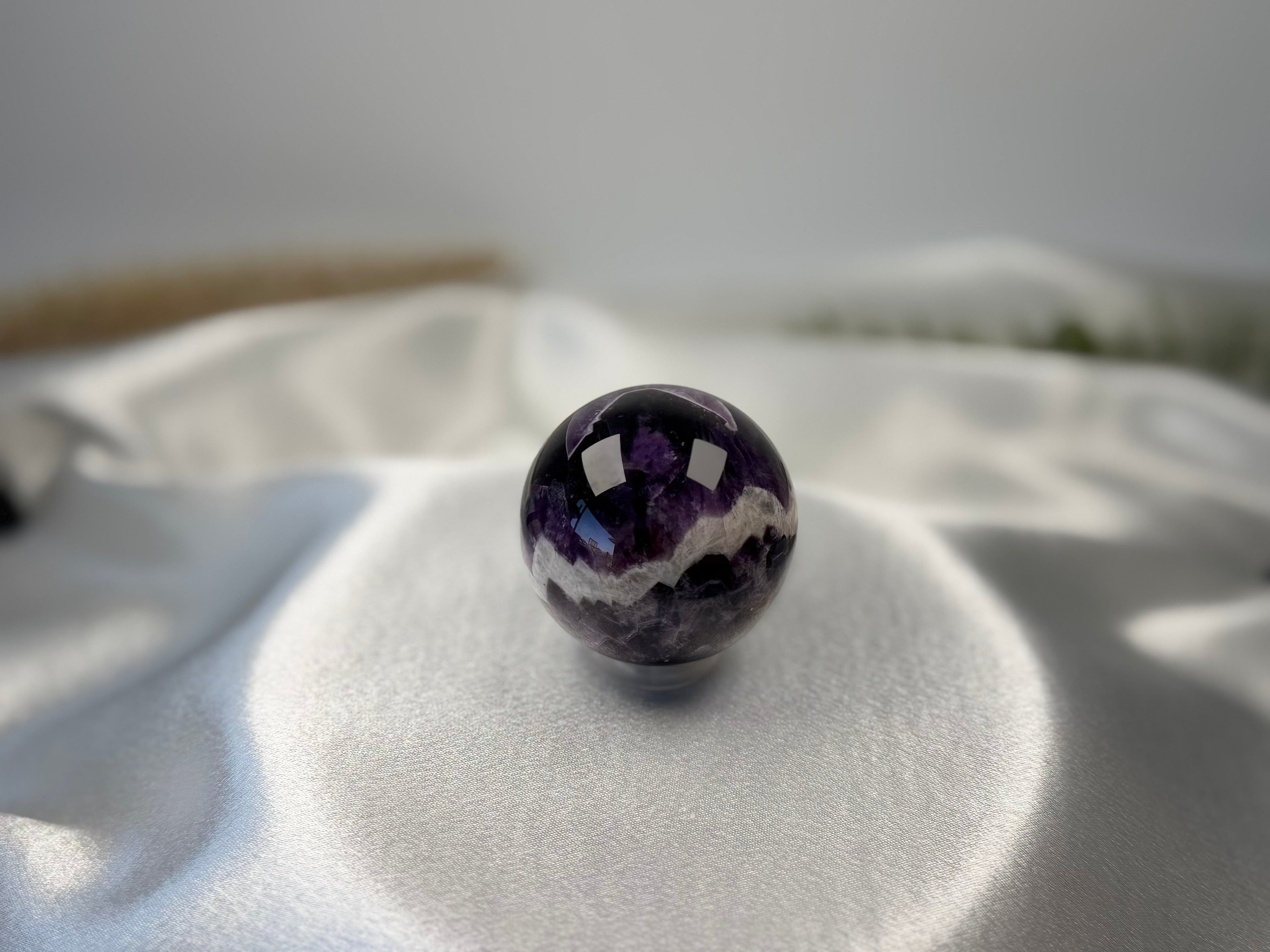 Treasured Energies polished chevron amethyst crystal sphere with deep purple and white V-shaped bands on satin fabric.