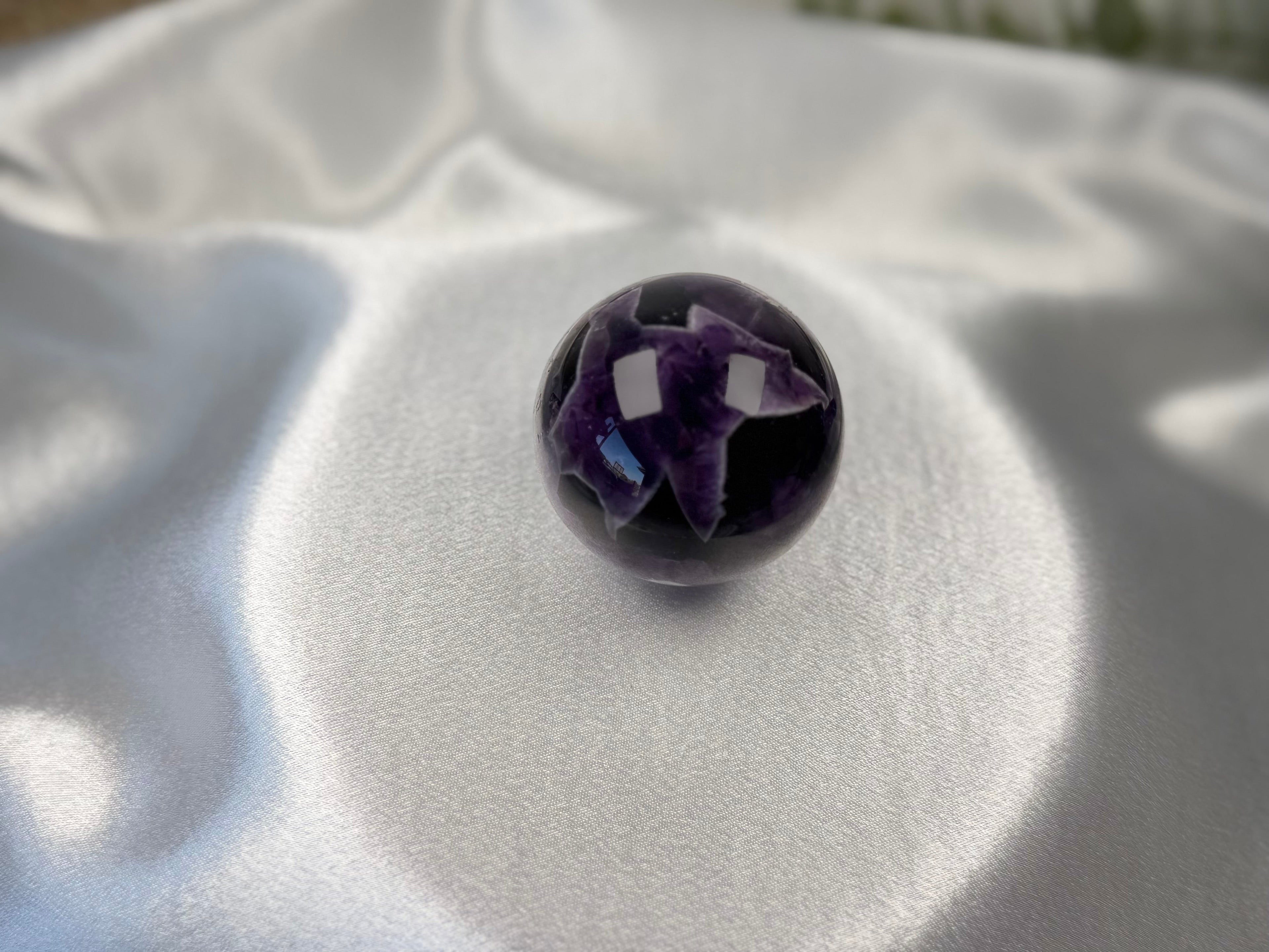 Treasured Energies polished Chevron Amethyst crystal sphere with deep purple and white V-shaped bands on satin fabric.