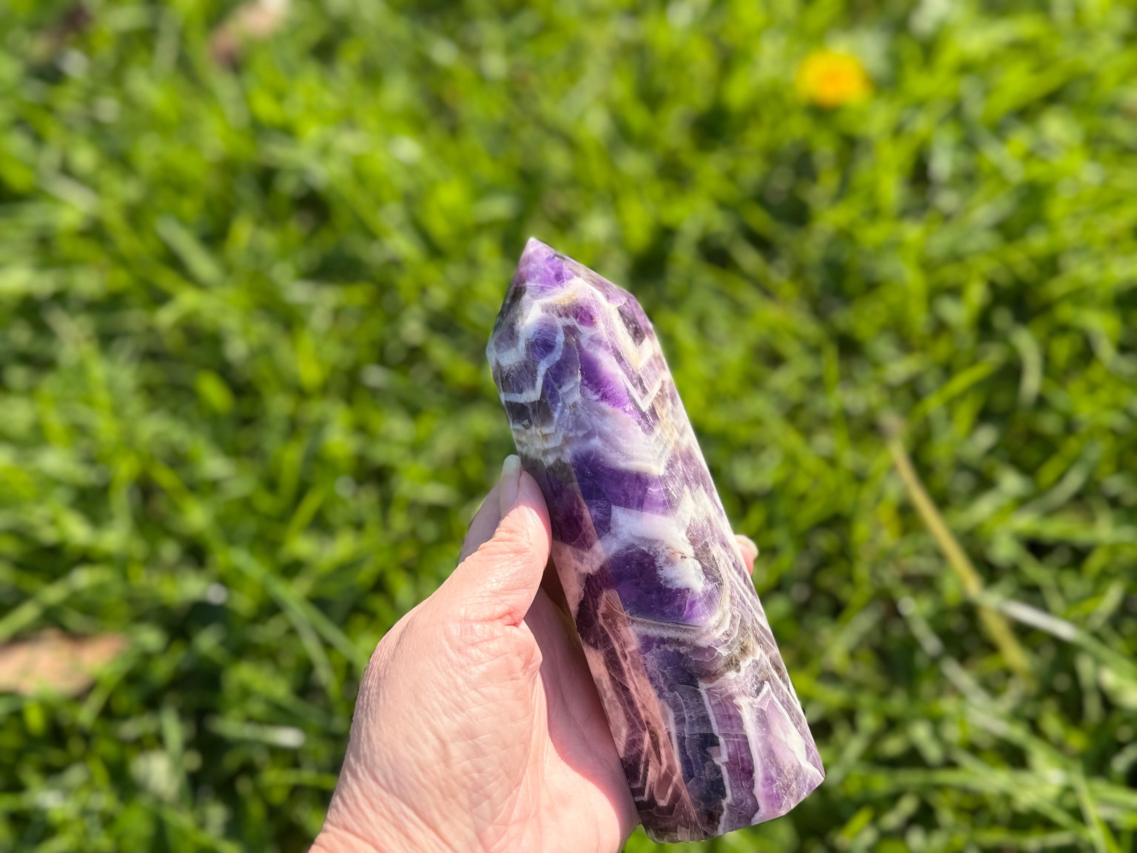 Treasured Energies Chevron Amethyst crystal tower held in hand against a blurred green grass background.