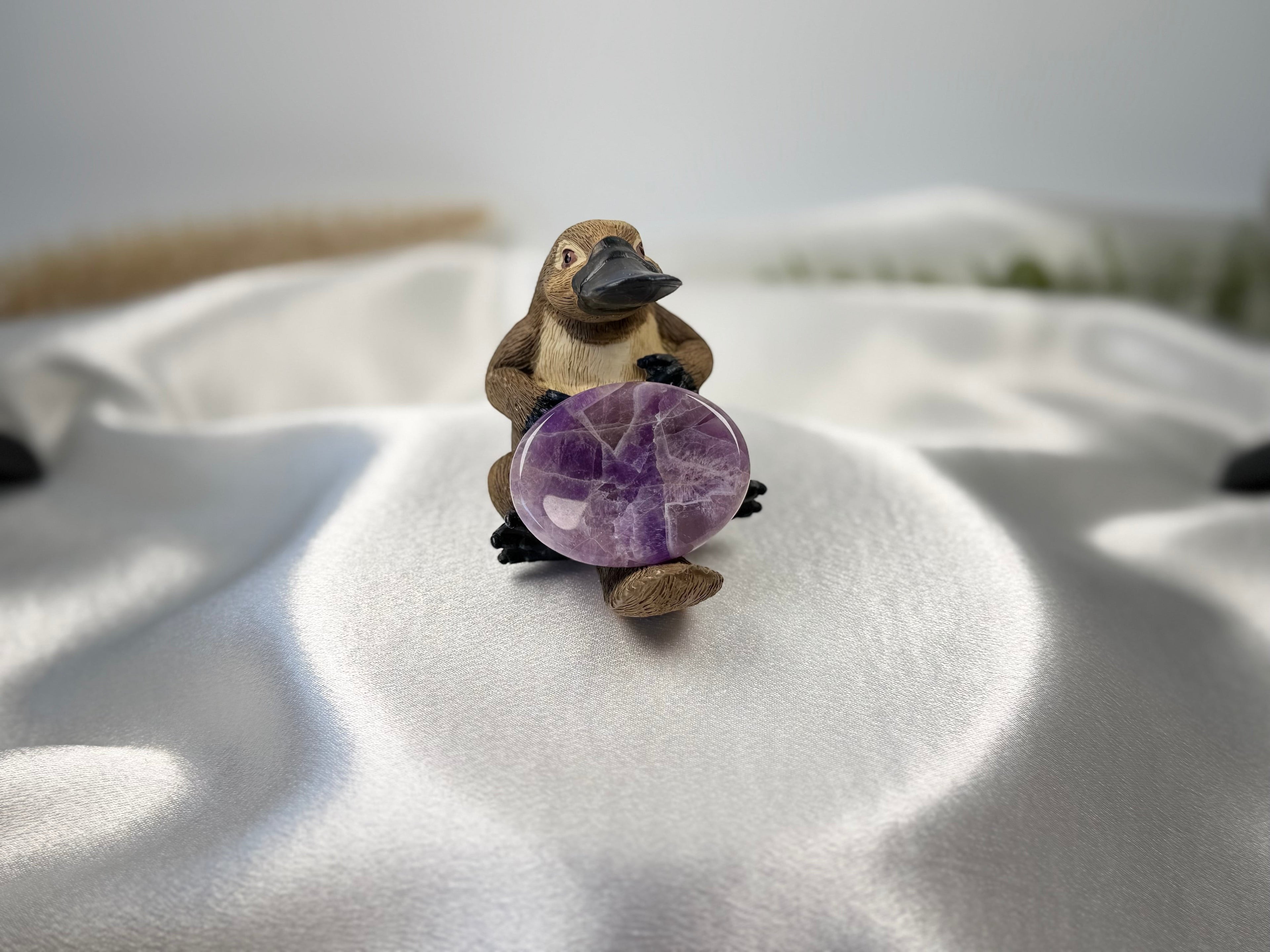 Treasured Energies Chevron Amethyst worry stone held by a small platypus figurine on a white satin fabric background.