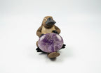 Treasured Energies Chevron Amethyst worry stone with purple and white bands held by a small platypus figurine.