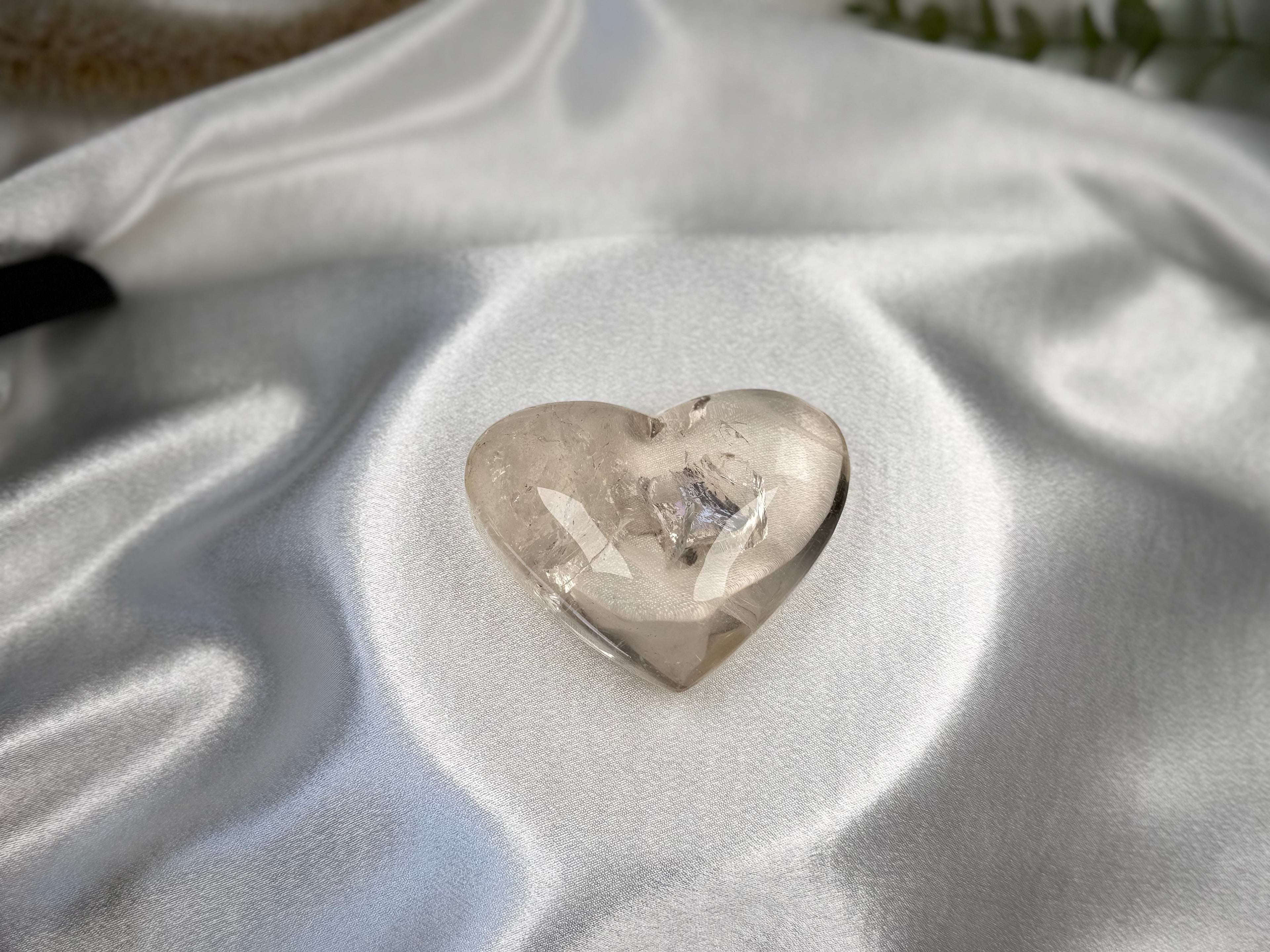 Treasured Energies clear quartz heart crystal with visible internal phantom on a white satin fabric background.