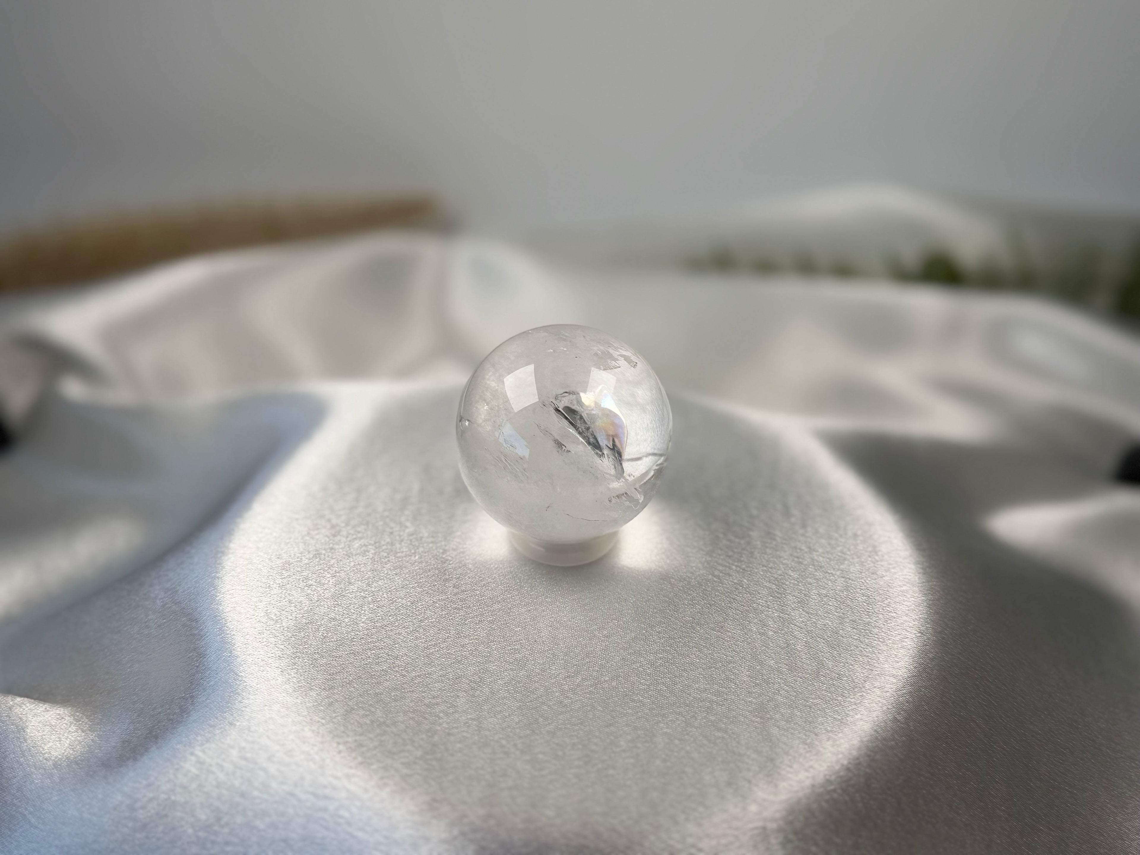 Treasured Energies clear quartz sphere with visible phantom inclusions resting on a smooth white satin fabric.