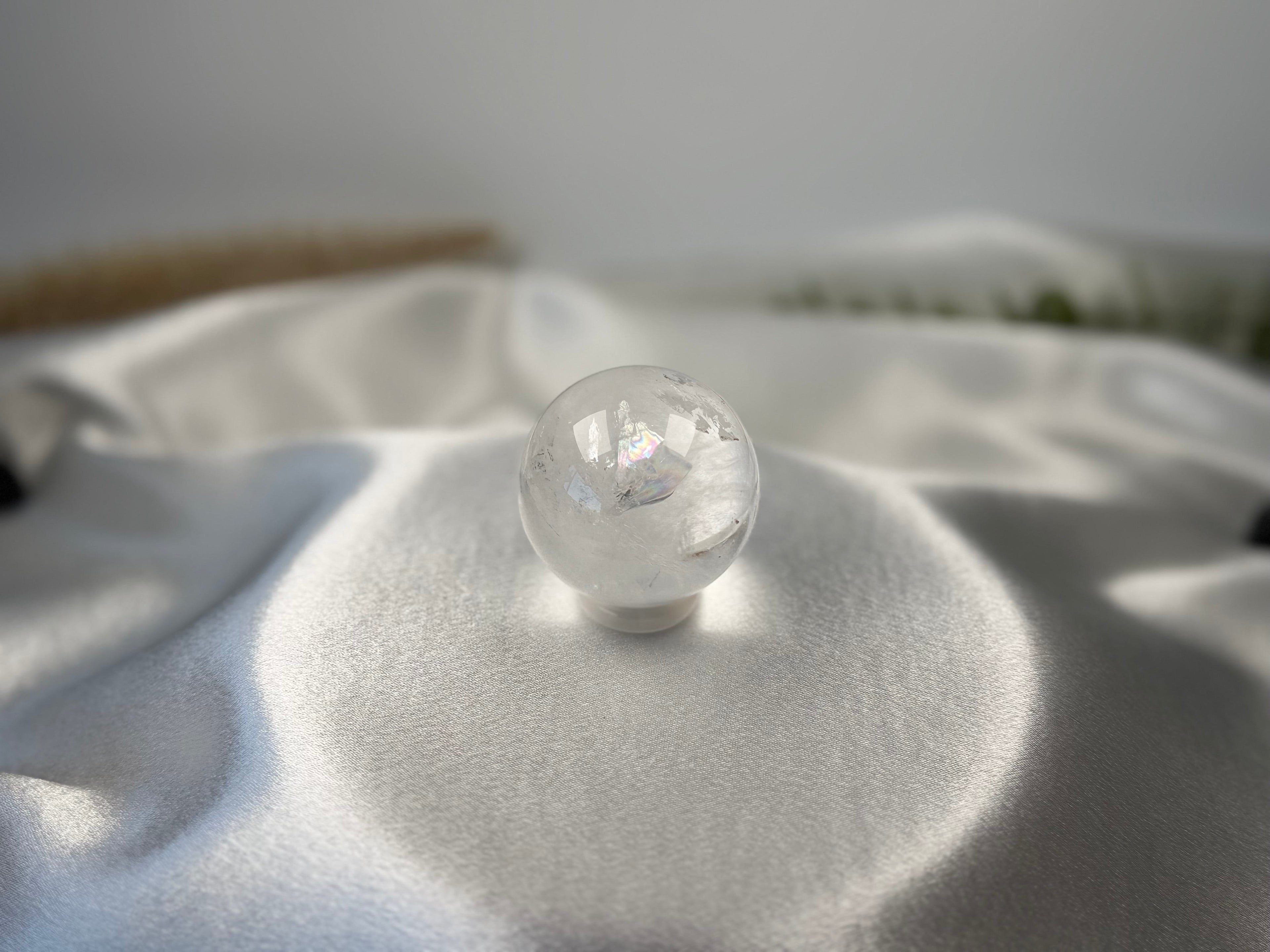 Treasured Energies clear quartz crystal sphere with visible phantom inclusions resting on white satin fabric.