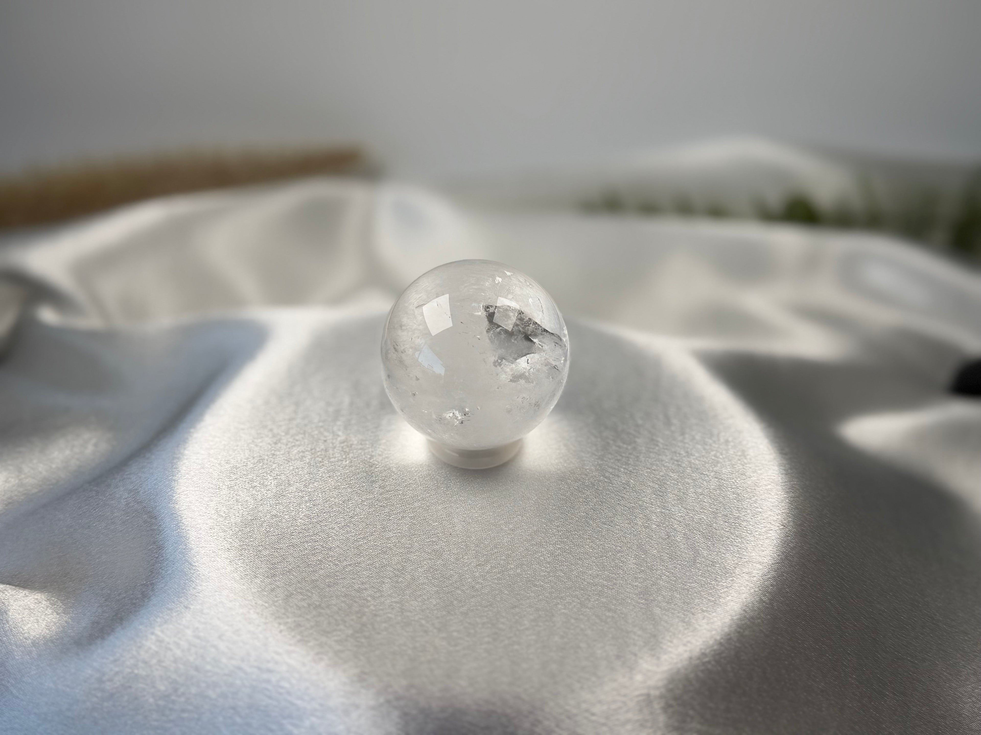 Treasured Energies clear quartz crystal sphere with visible phantom inclusions resting on white satin fabric.