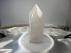 Treasured Energies clear quartz crystal tower with a polished point and raw base on a white satin fabric background.