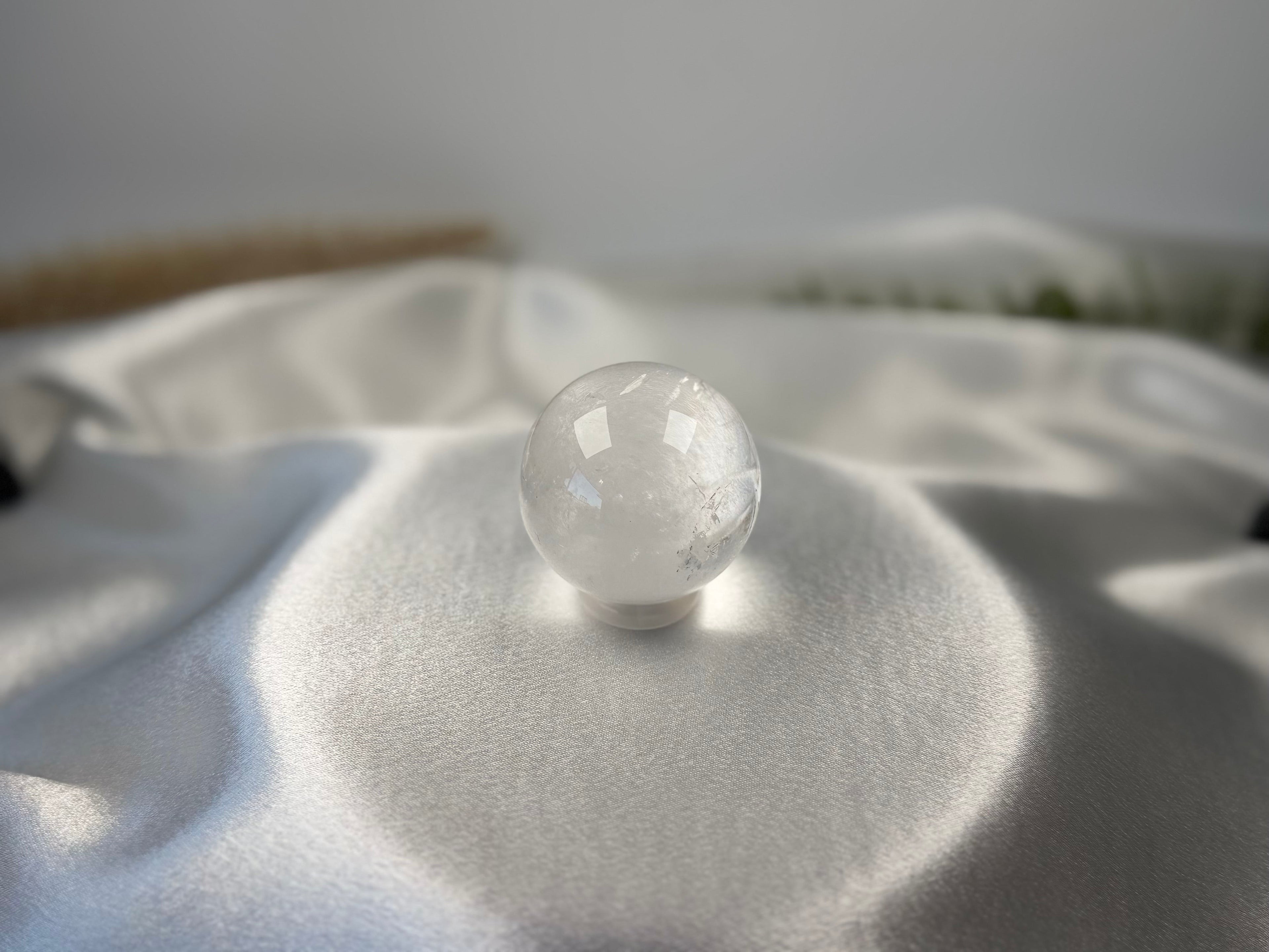 Treasured Energies clear quartz crystal sphere with visible phantom inclusions resting on a smooth white satin fabric.