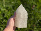 Treasured Energies clear quartz crystal tower with a raw base and polished point held against a green outdoor background