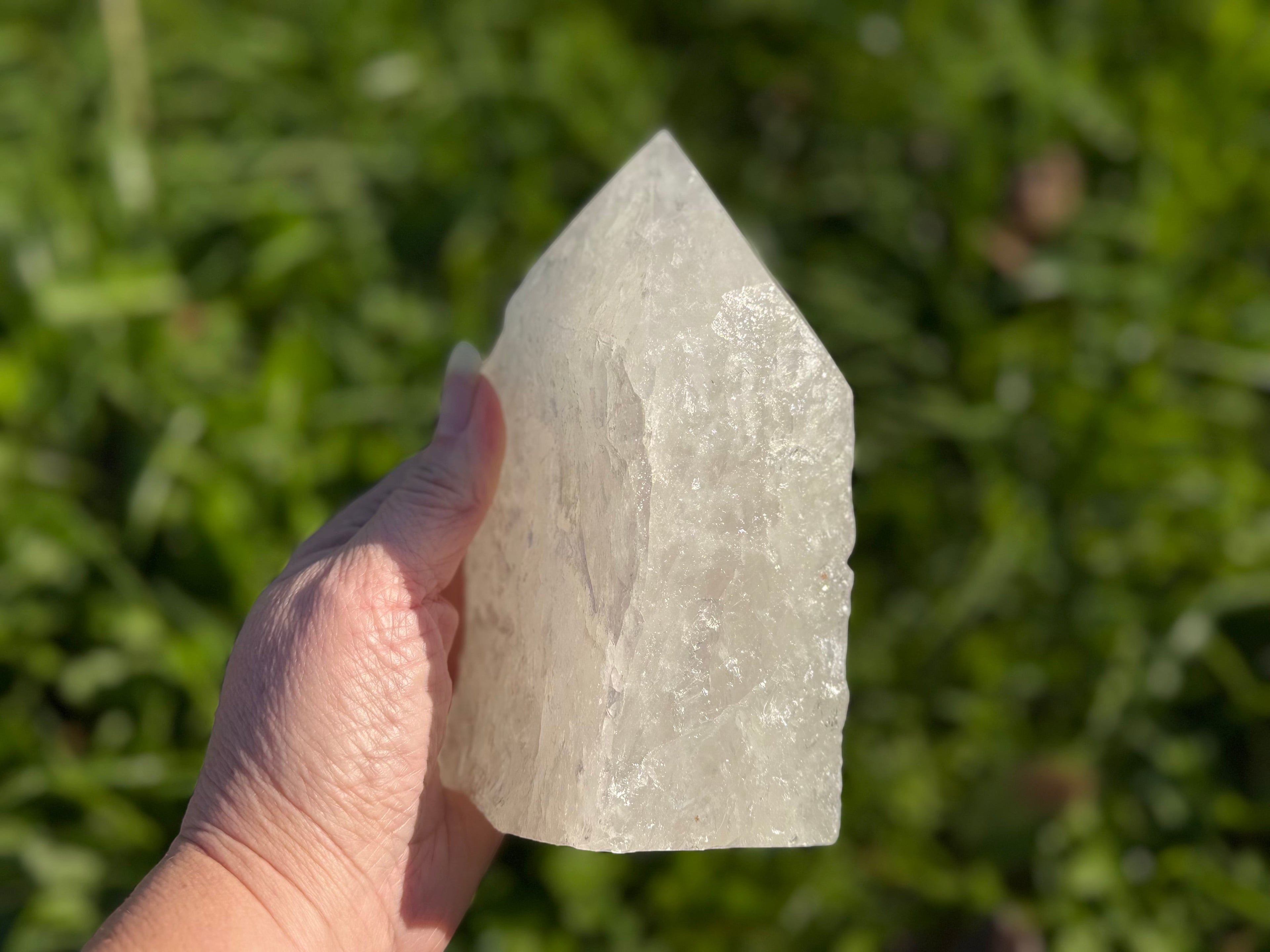 Treasured Energies clear quartz crystal tower with a raw base and polished point held against a green outdoor background