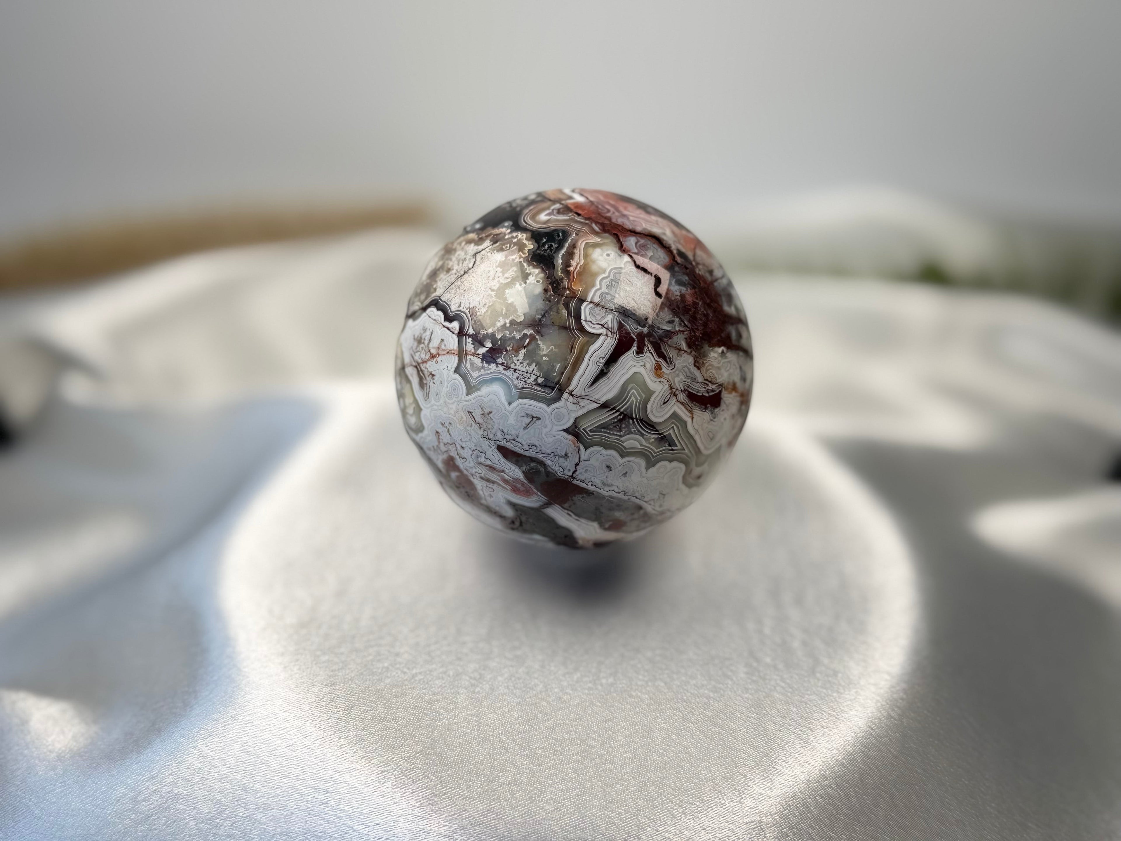 Treasured Energies polished Crazy Lace Agate crystal sphere with intricate swirling patterns on white satin fabric.
