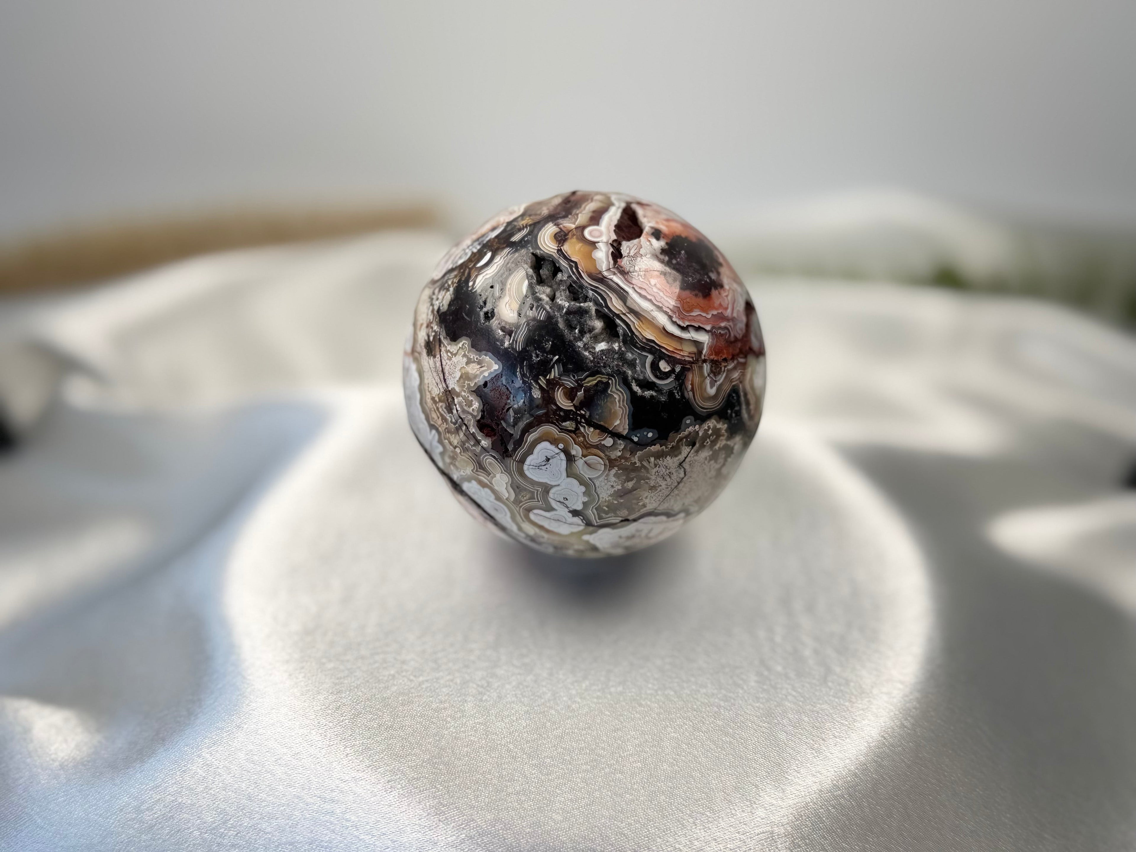 Treasured Energies polished Crazy Lace Agate crystal sphere with intricate swirling patterns on a white silk cloth.