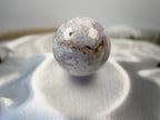 Treasured Energies flower agate crystal sphere with natural druzy inclusions resting on a soft white satin fabric.