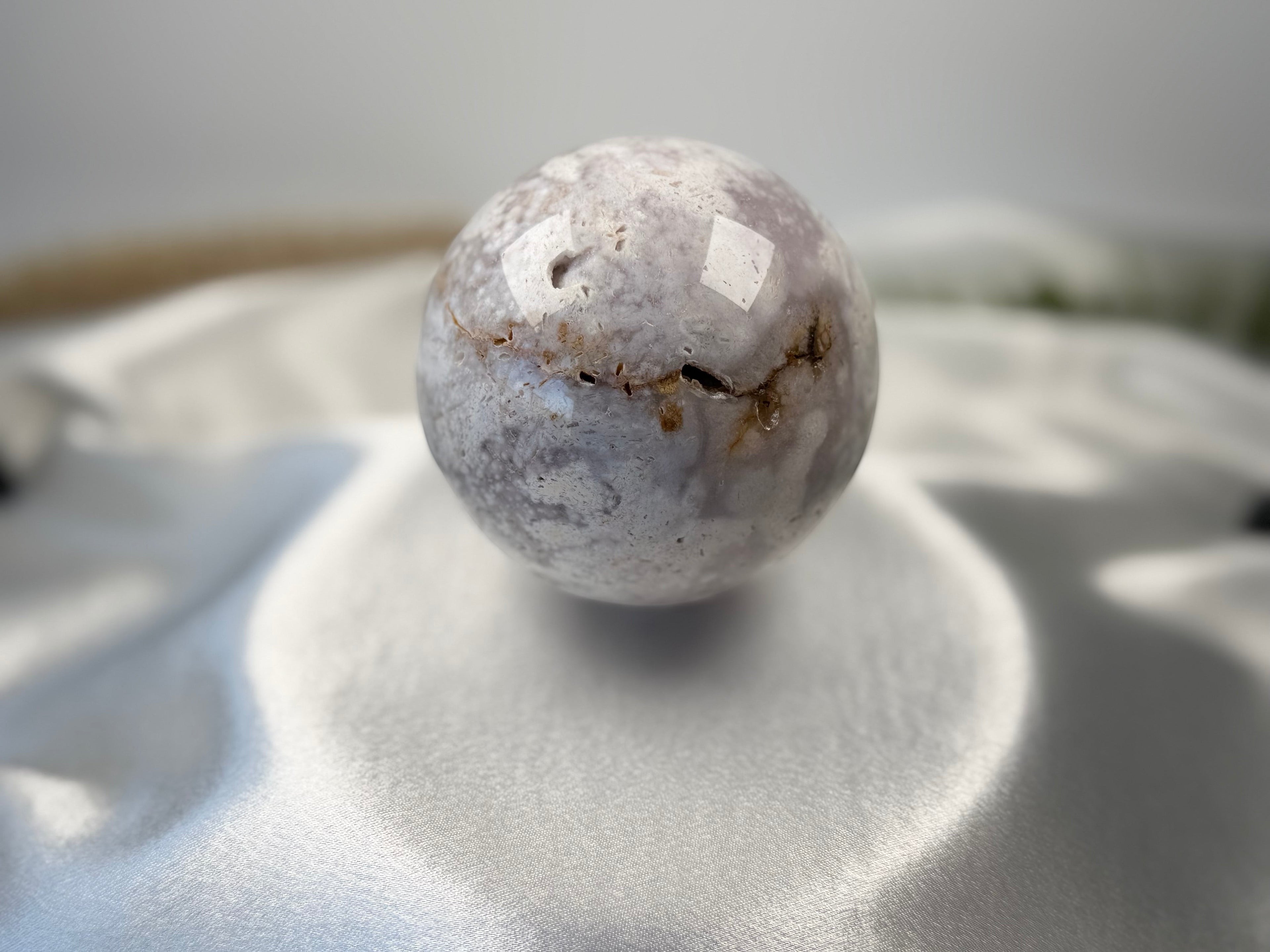 Treasured Energies flower agate crystal sphere with natural druzy inclusions resting on a soft white satin fabric.