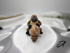 Treasured Energies flower agate crystal heart held by a small platypus figurine on a white satin fabric background.