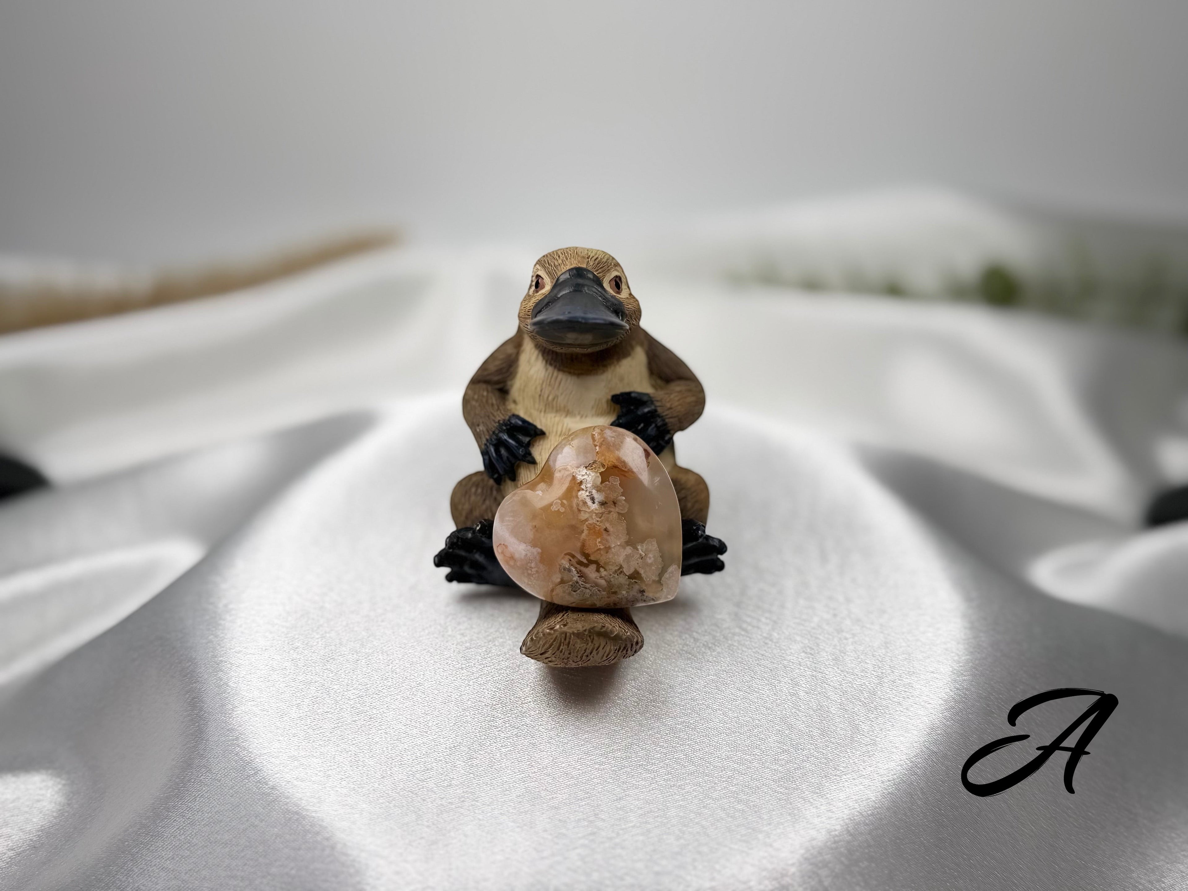 Treasured Energies flower agate crystal heart held by a small platypus figurine on a white satin fabric background.