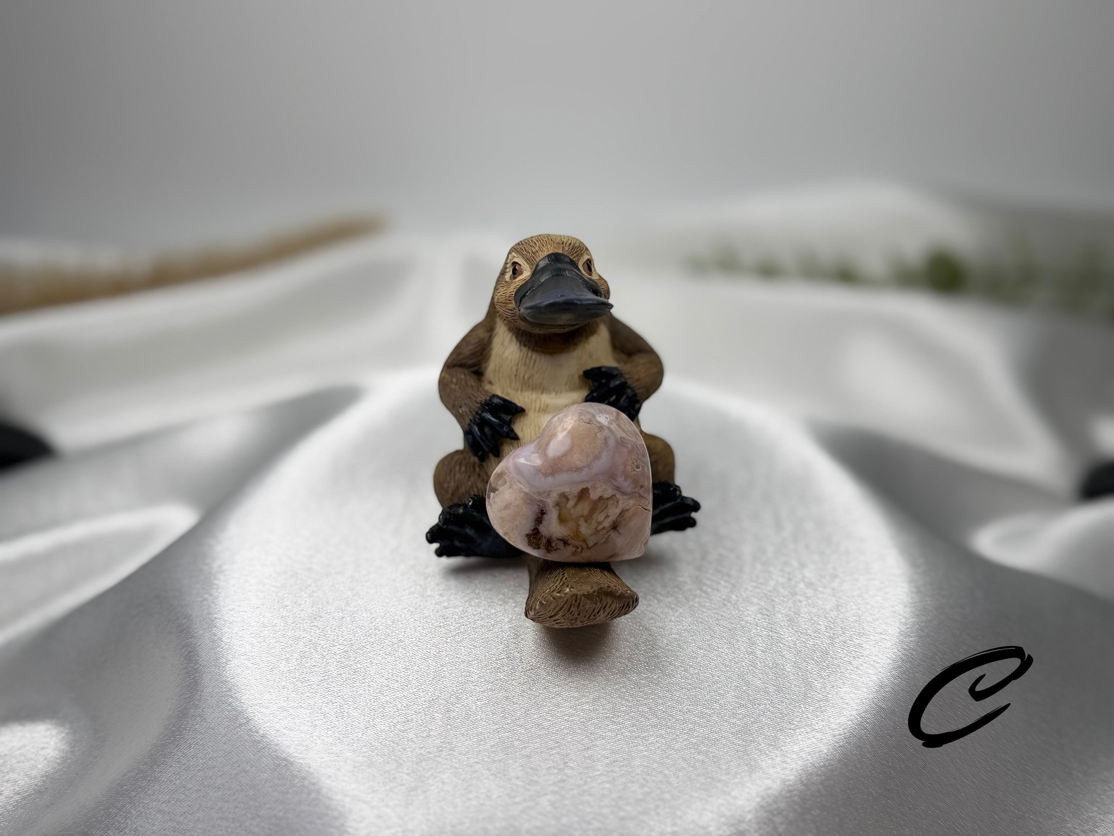 Treasured Energies flower agate crystal heart held by a small platypus figurine on a white satin background.