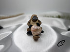 Treasured Energies polished flower agate crystal heart held by a small brown platypus figurine on white satin fabric.