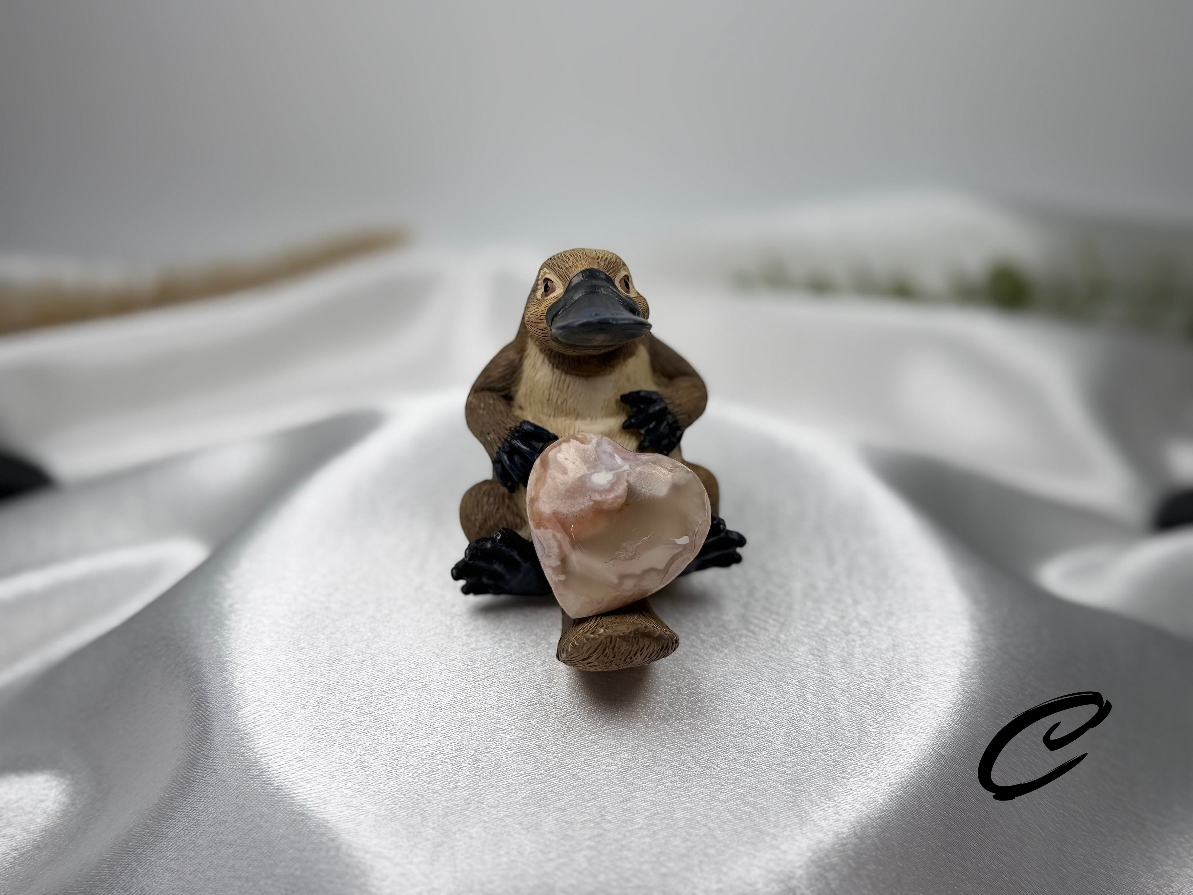 Treasured Energies polished flower agate crystal heart held by a small brown platypus figurine on white satin fabric.