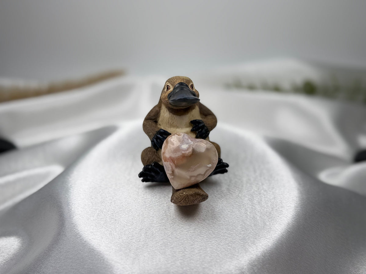 Treasured Energies polished flower agate crystal heart held by a small brown platypus figurine on white satin fabric.