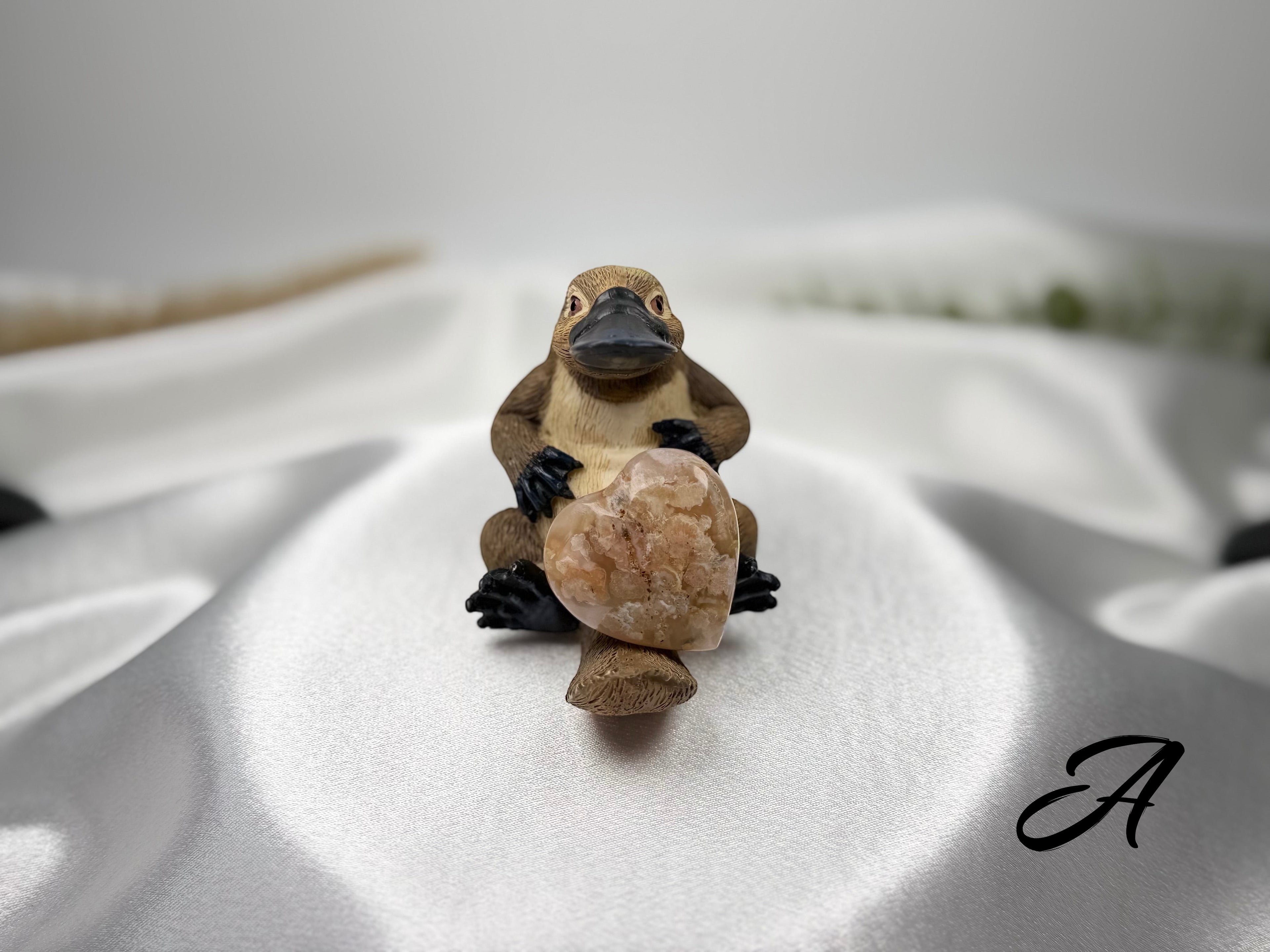 Treasured Energies flower agate crystal heart held by a small platypus figurine on a white satin background.