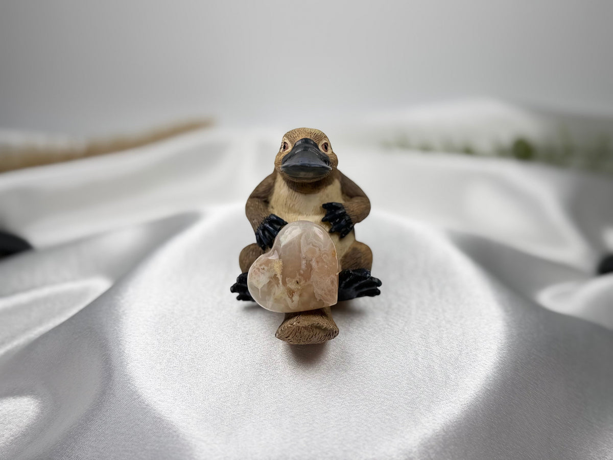 Treasured Energies flower agate crystal heart held by a small platypus figurine on a white satin background.