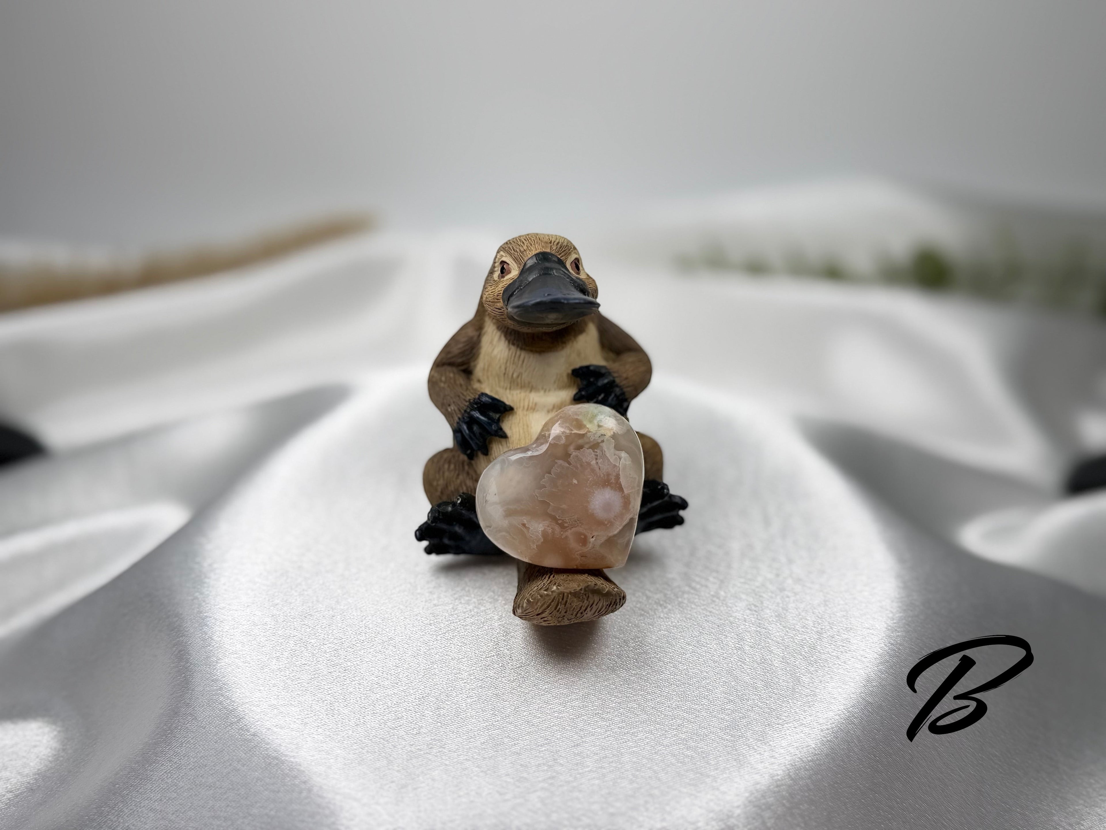 Treasured Energies flower agate crystal heart held by a small platypus figurine on a white satin background