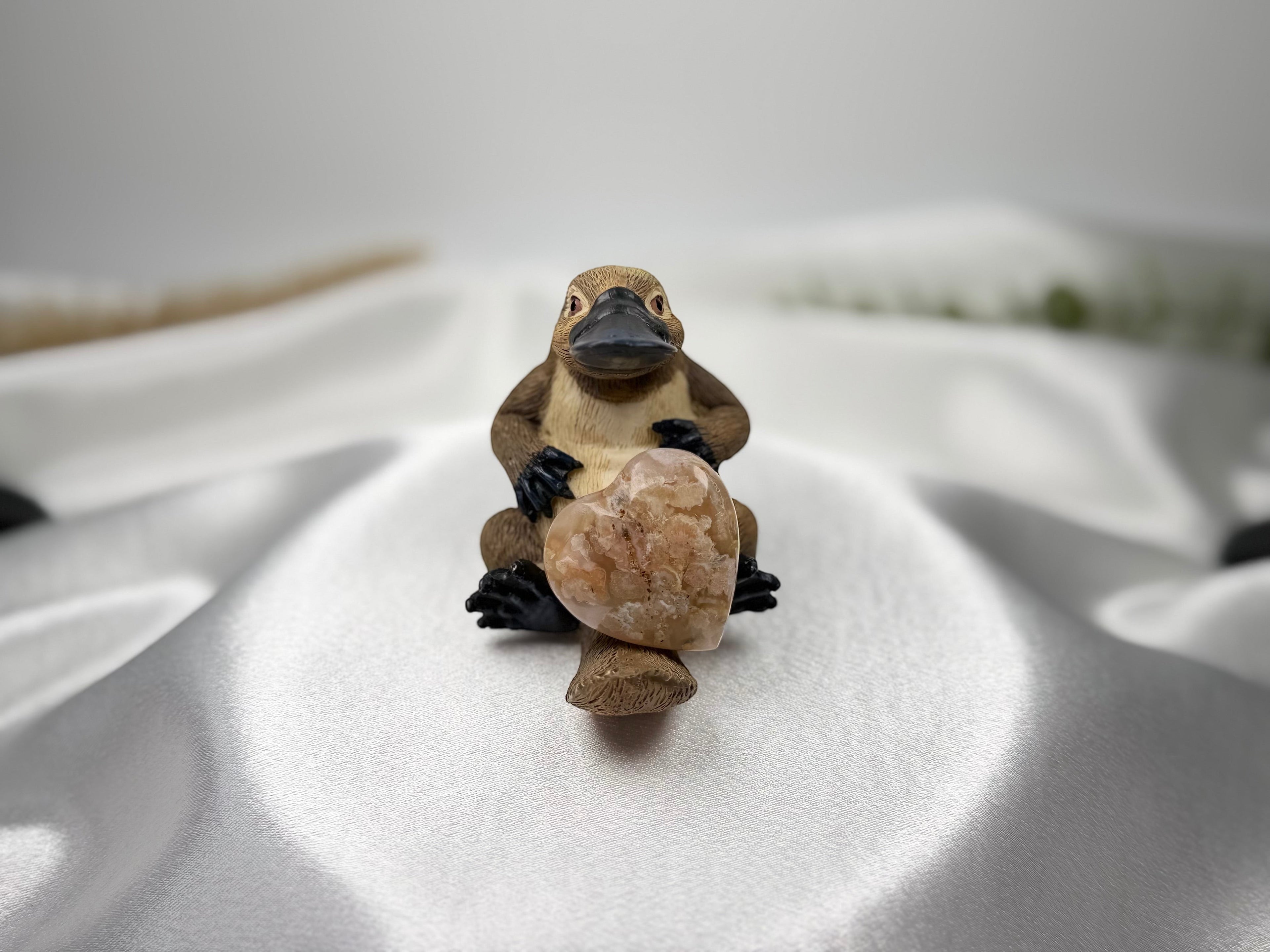 Treasured Energies flower agate crystal heart held by a small platypus figurine on a white satin background.