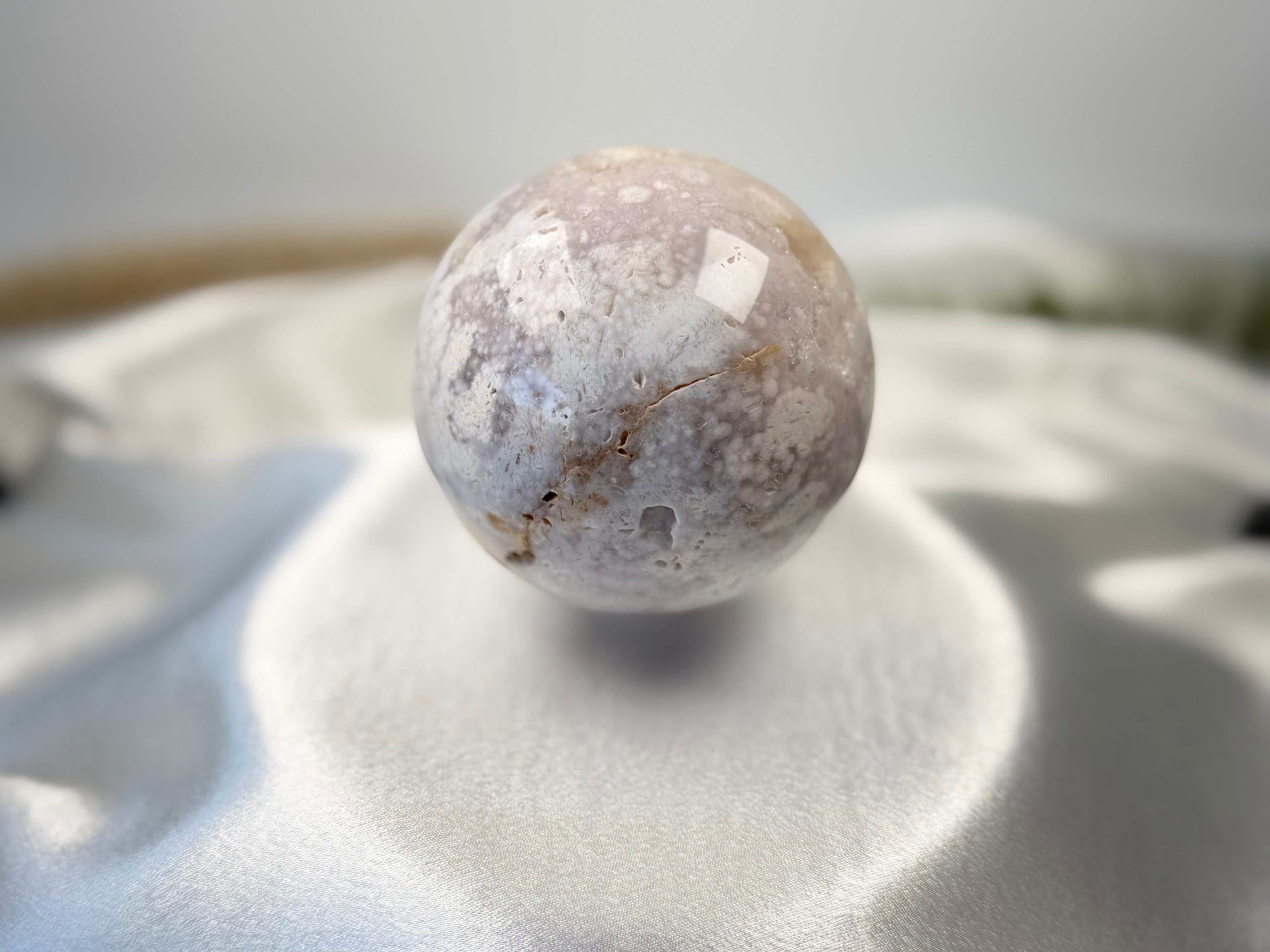 Treasured Energies flower agate sphere with druzy inclusions resting on a soft white satin fabric