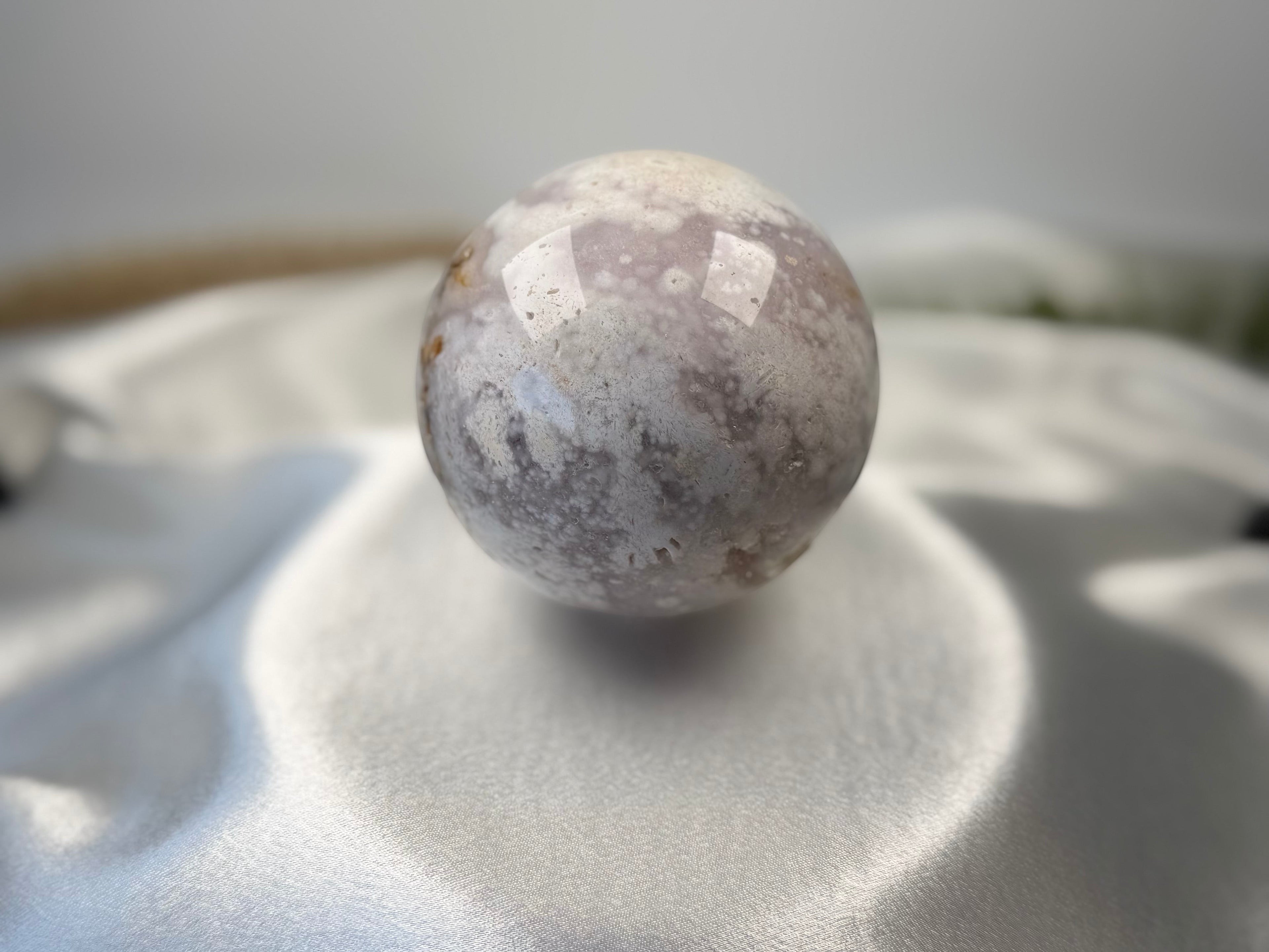 Treasured Energies flower agate sphere with druzy inclusions resting on a soft white satin fabric