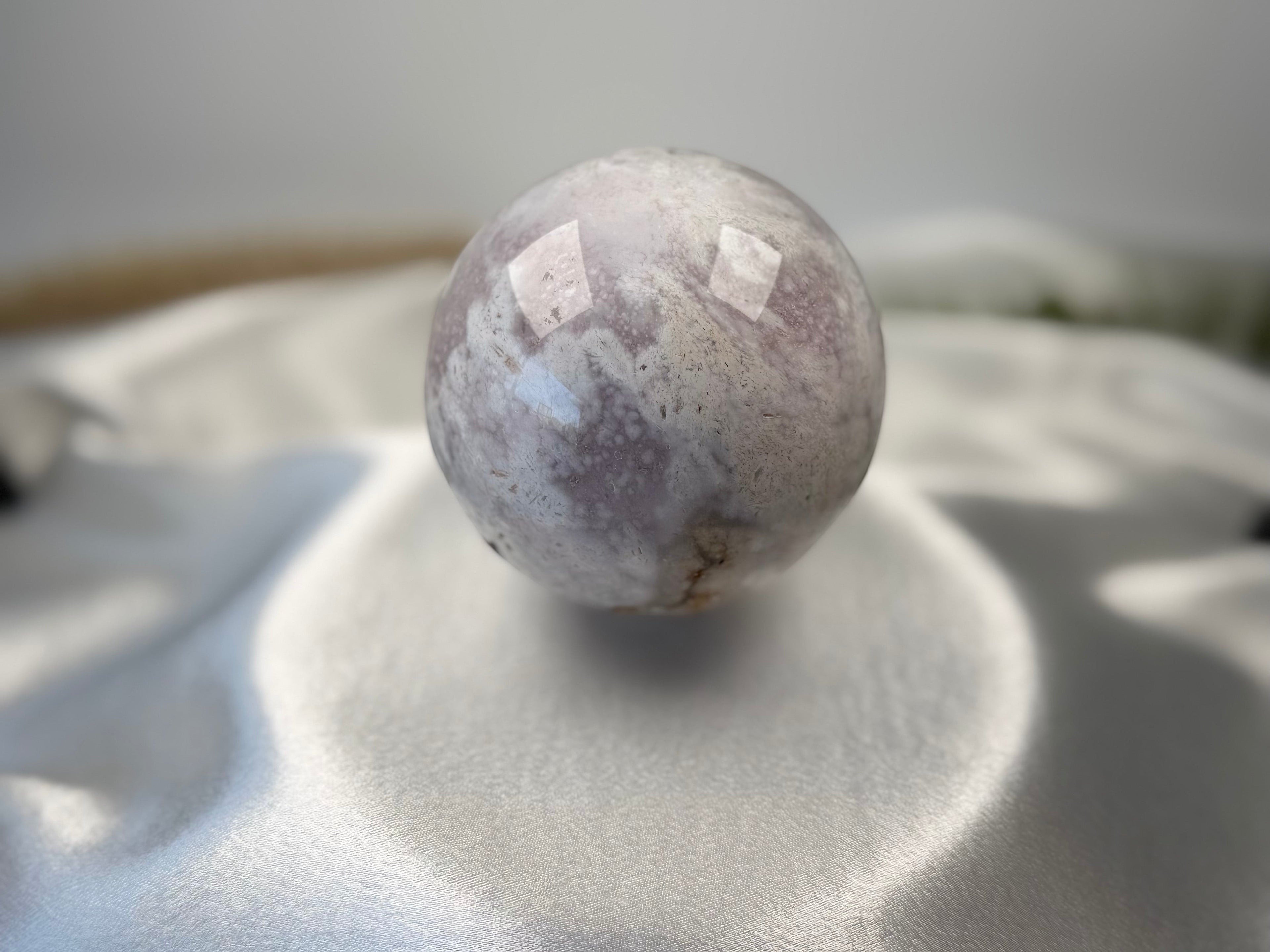 Treasured Energies polished flower agate crystal sphere with druzy inclusions resting on a soft white satin fabric.