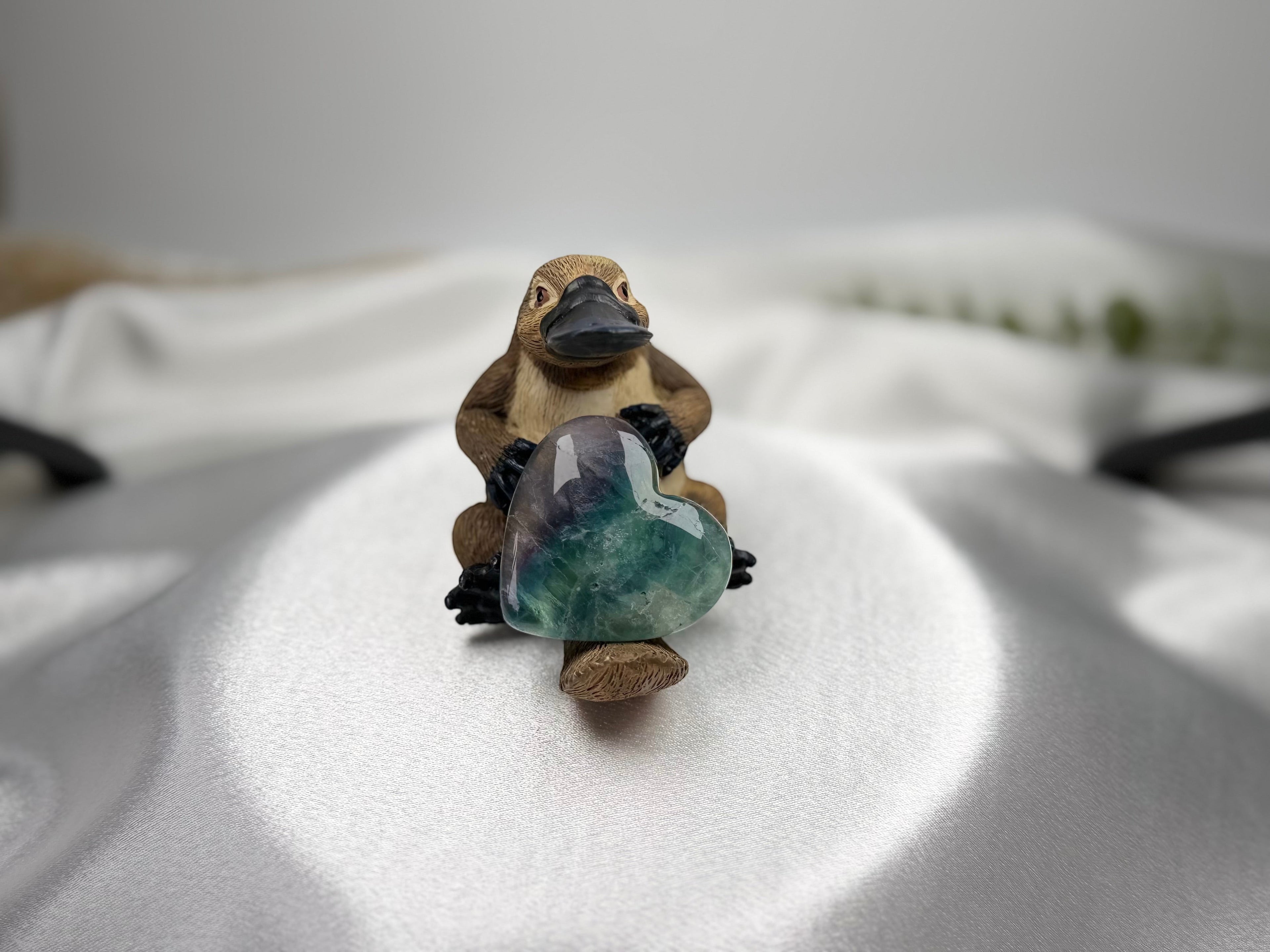 Treasured Energies polished fluorite crystal heart held by a small platypus figurine on a white satin background.