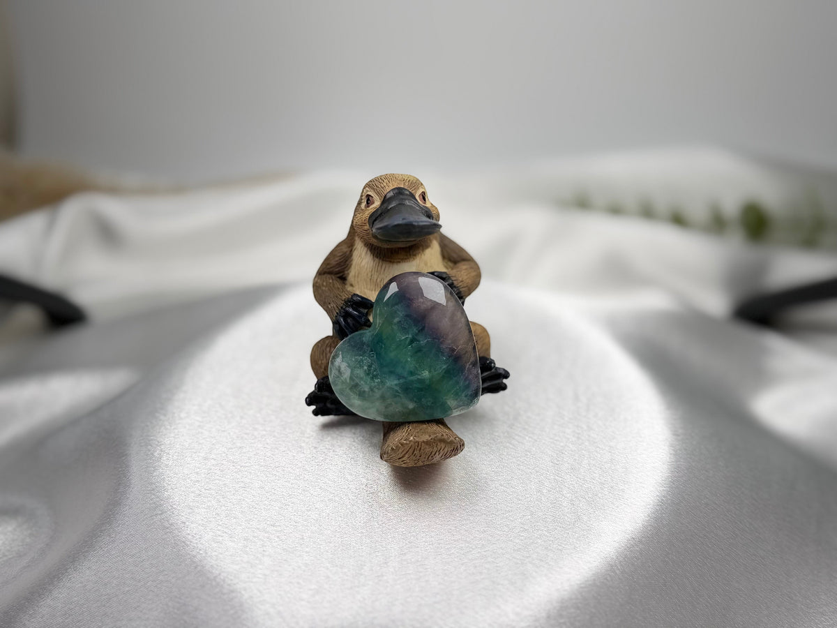 Treasured Energies polished green and purple fluorite crystal heart held by a small platypus figurine on satin fabric.