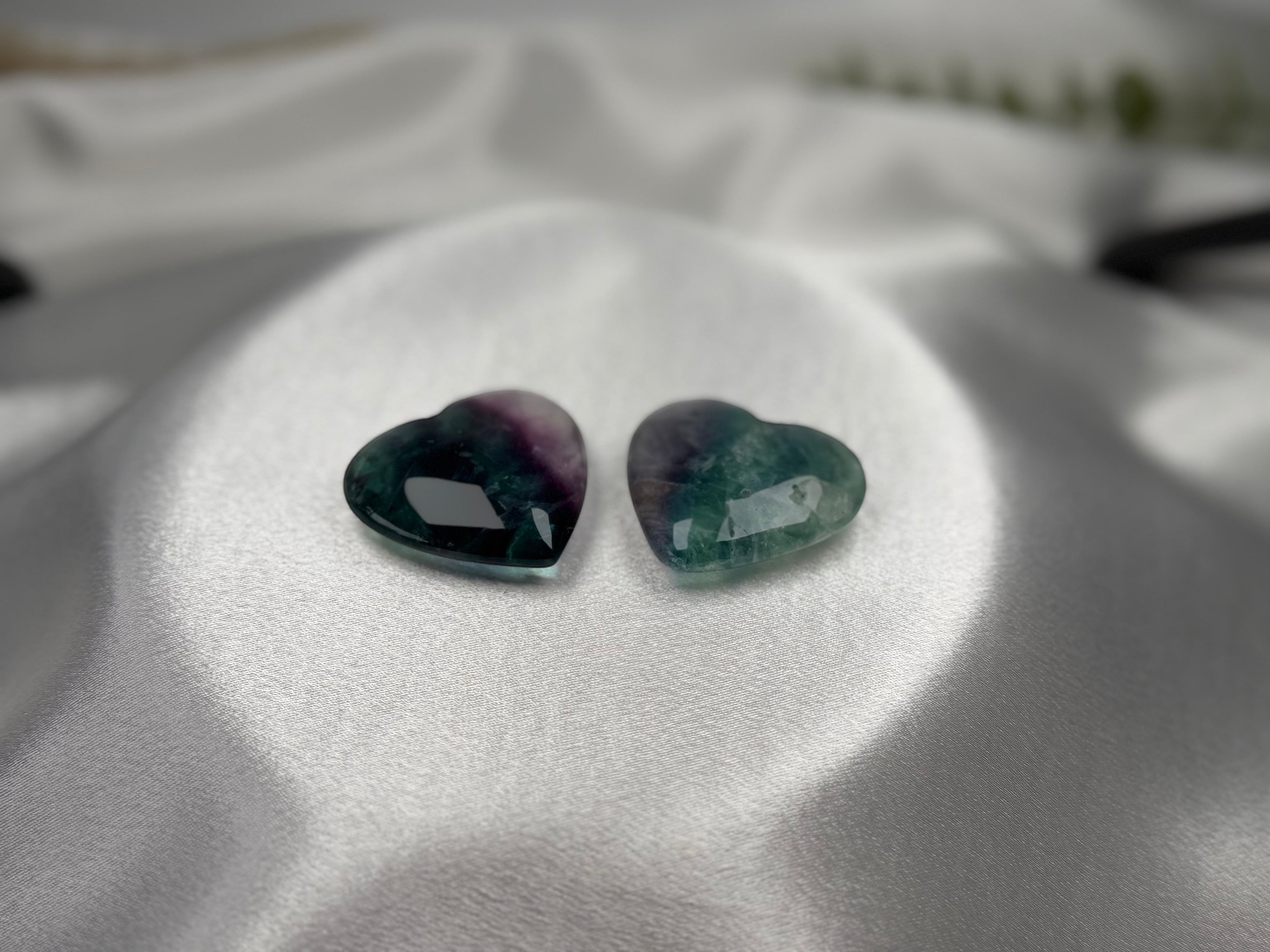 Treasured Energies polished fluorite crystal hearts in purple and green hues resting on a smooth white satin fabric.