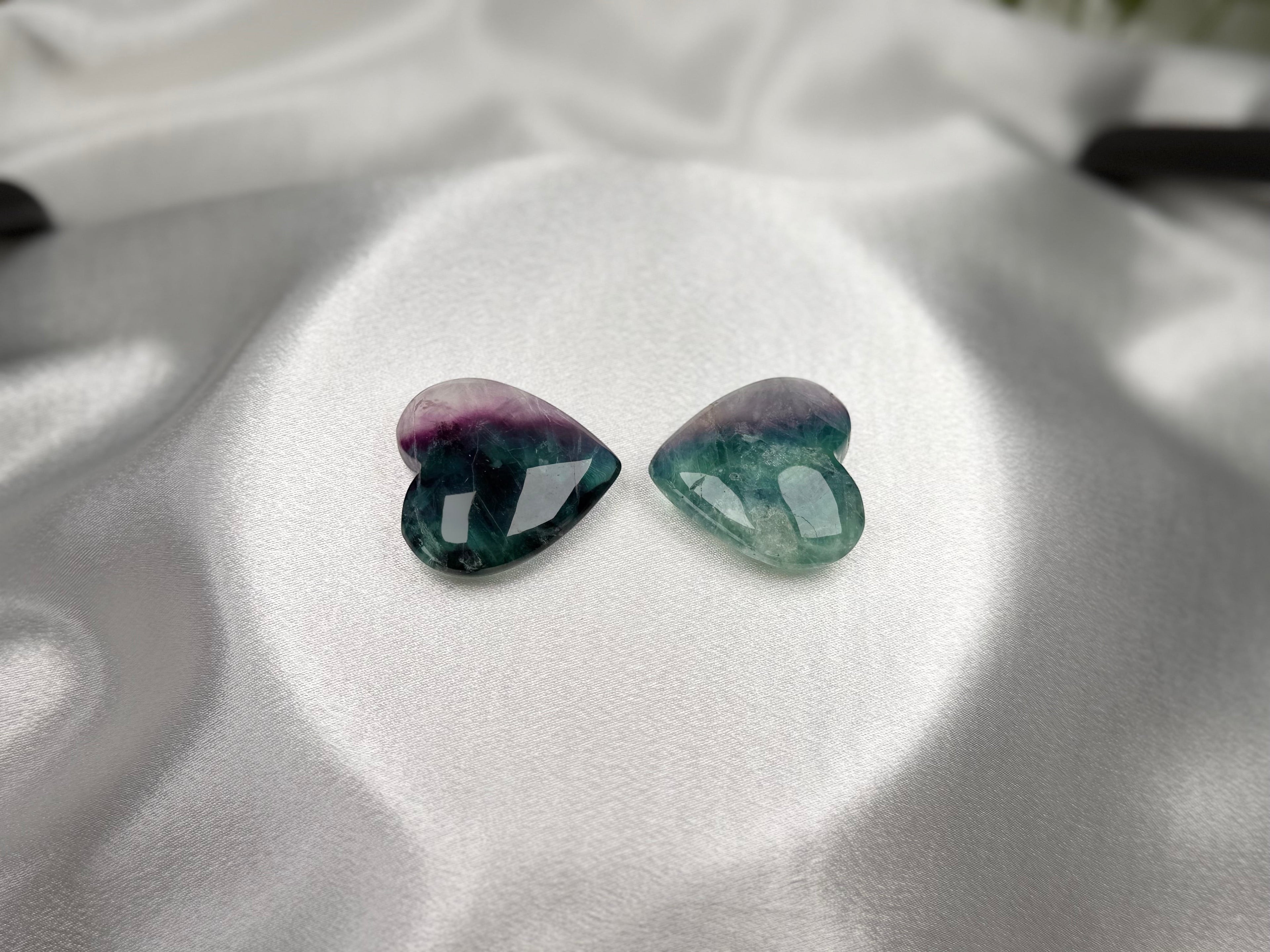 Treasured Energies polished fluorite crystal hearts in purple and green hues resting on a white satin fabric background.