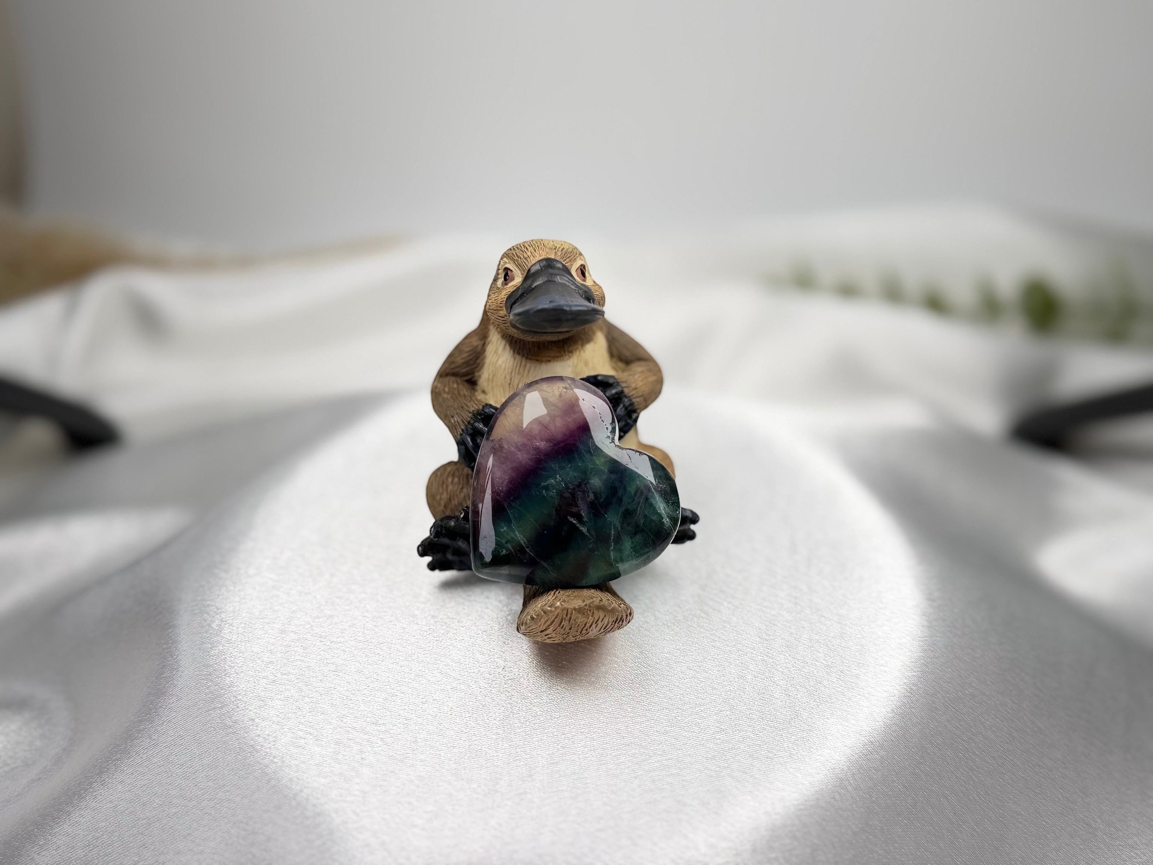 Treasured Energies polished heart-shaped fluorite crystal held by a small platypus figurine on a white satin background.