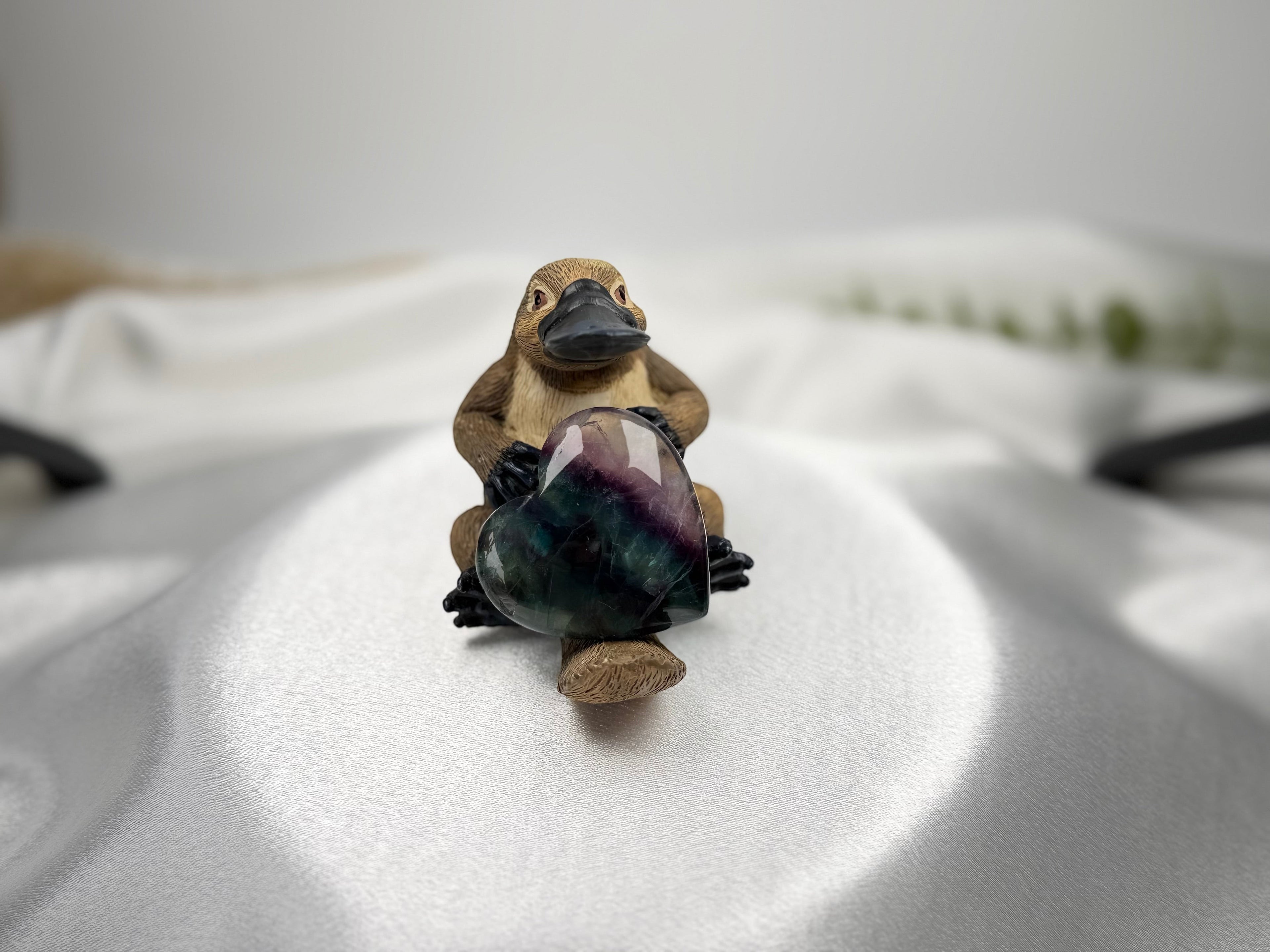 Treasured Energies polished heart-shaped fluorite crystal held by a small platypus figurine on white satin fabric.