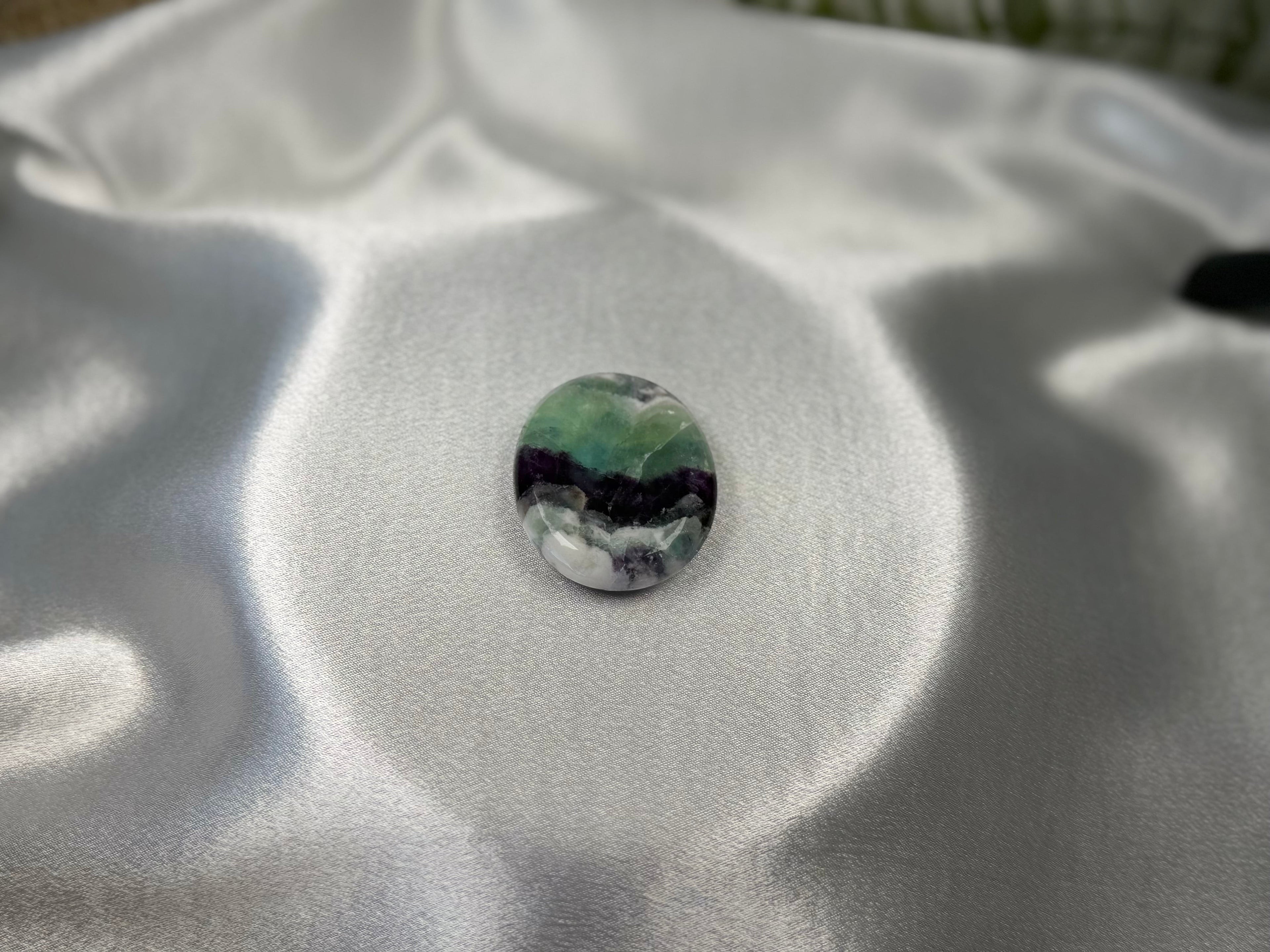 Treasured Energies smooth, oval-shaped fluorite worry stone with purple and green bands on a white satin background.