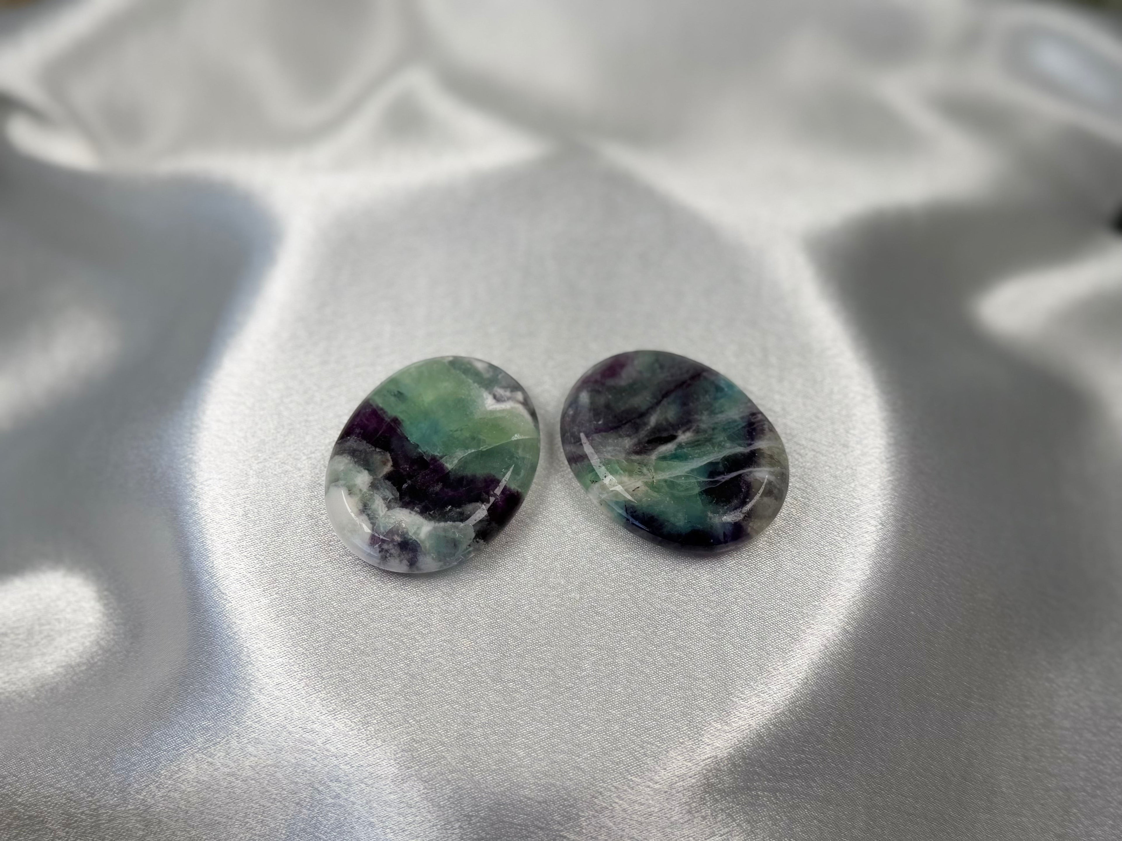 Two smooth, oval-shaped Treasured Energies fluorite worry stones with purple and green banding on a satin fabric.