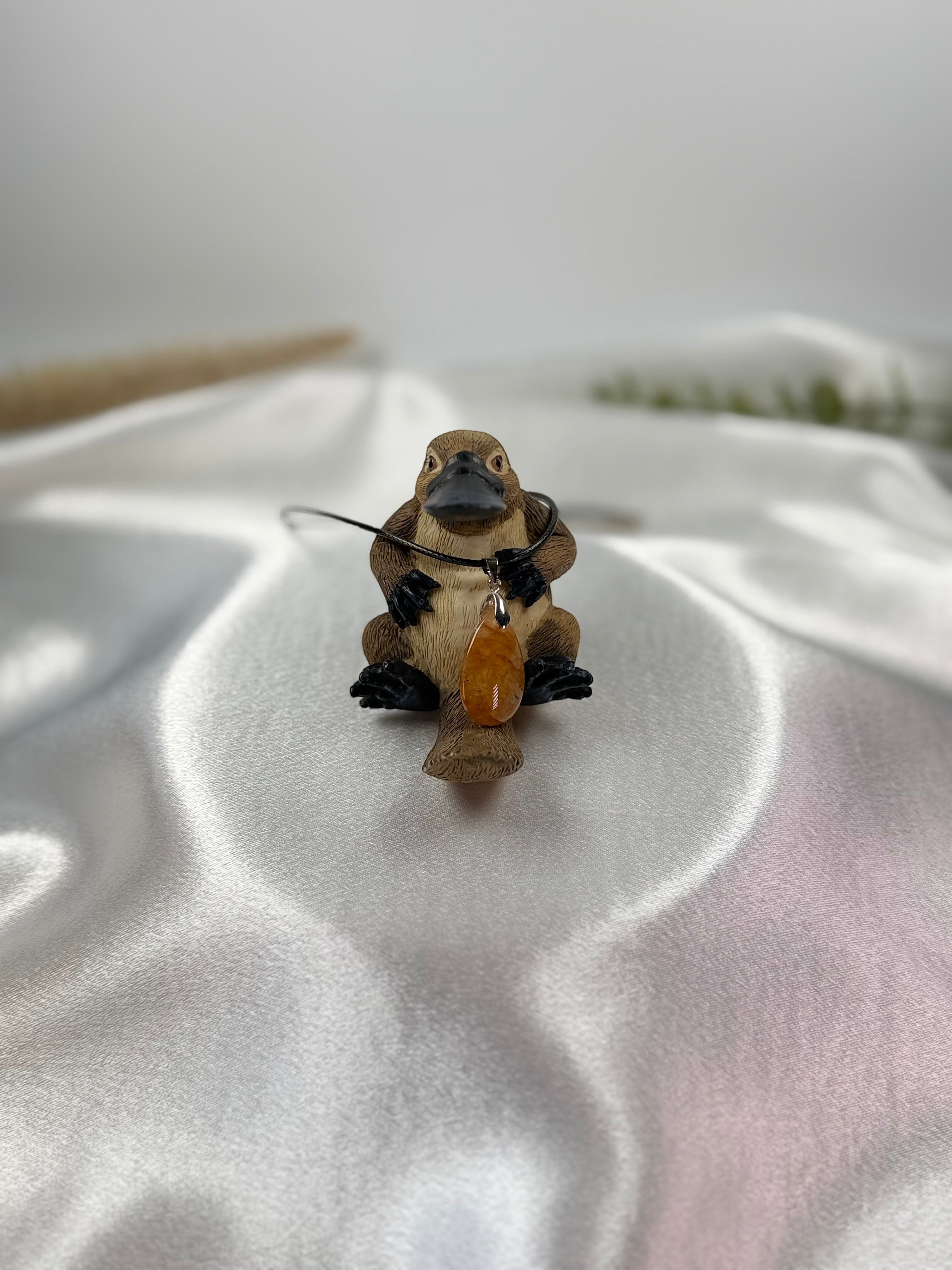 Treasured Energies golden healer quartz crystal pendant on a black cord, displayed on a platypus figurine.