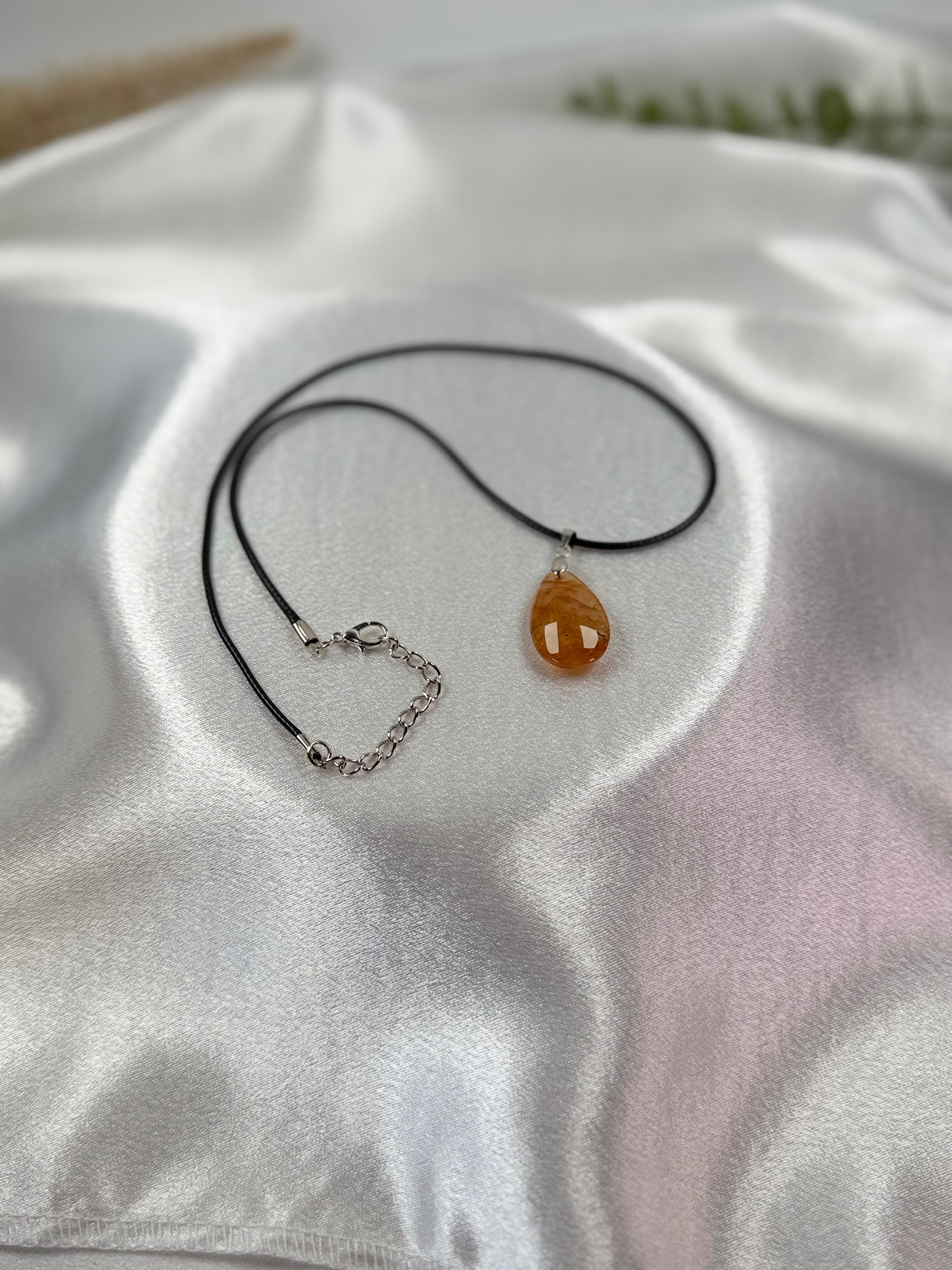 Treasured Energies golden healer quartz teardrop pendant on a black cord necklace resting on white satin fabric.