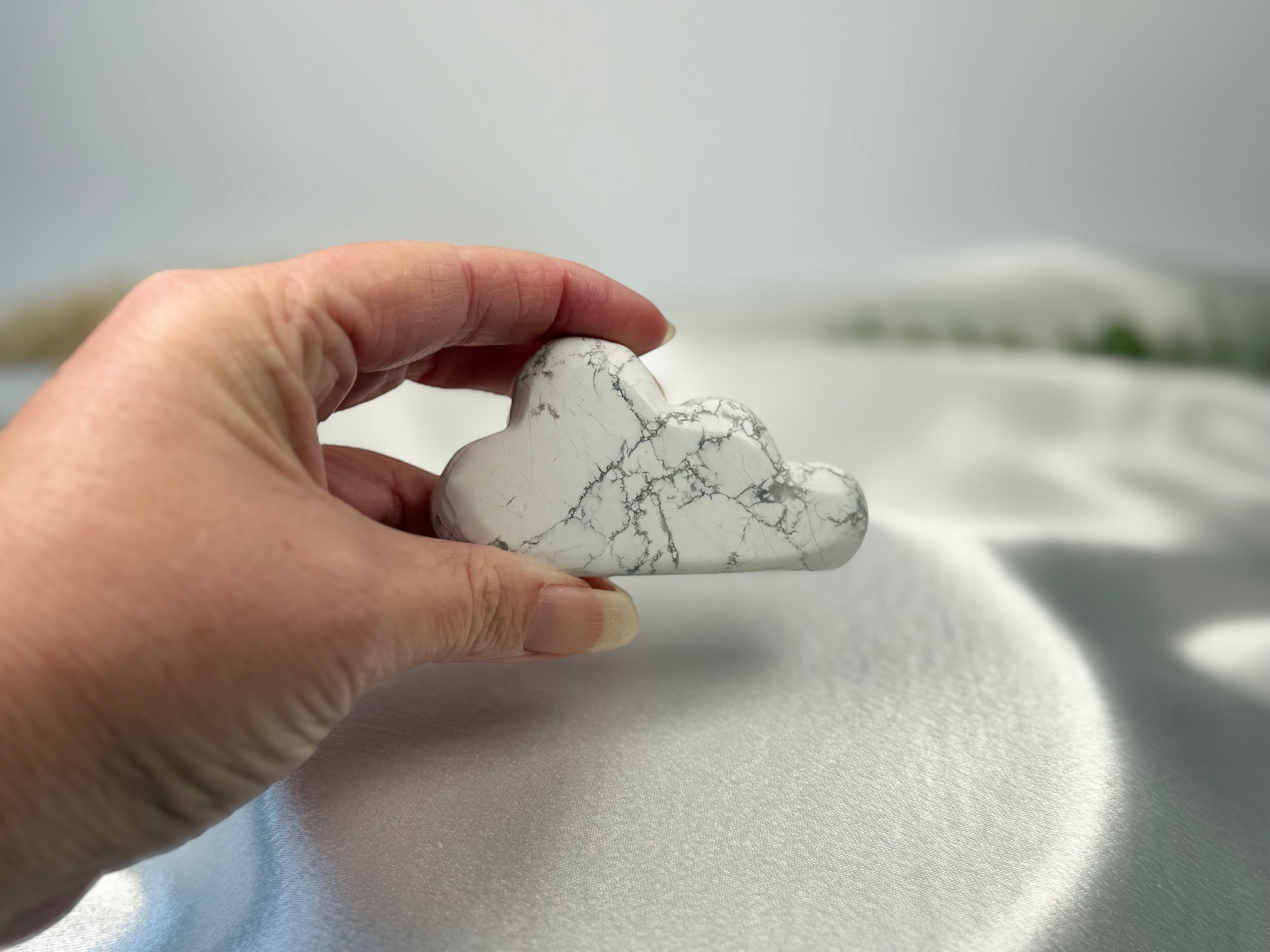 Treasured Energies hand carved howlite crystal cloud with grey veining held in a hand against a white satin background.