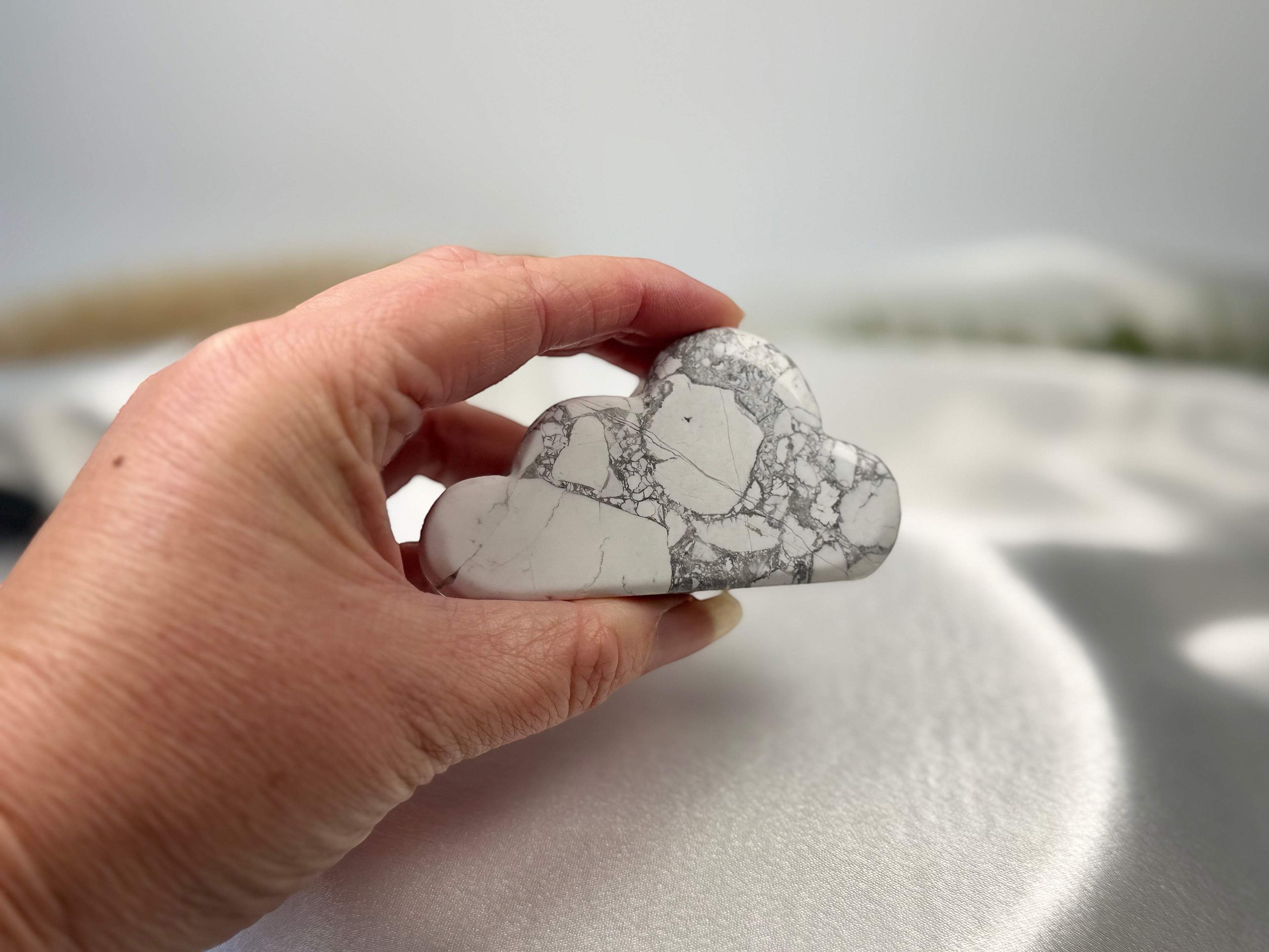 Treasured Energies hand carved howlite crystal cloud with grey veining held in a hand against a white satin background.