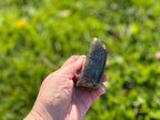 Treasured Energies polished labradorite freeform crystal held in a hand against a blurred green outdoor background.
