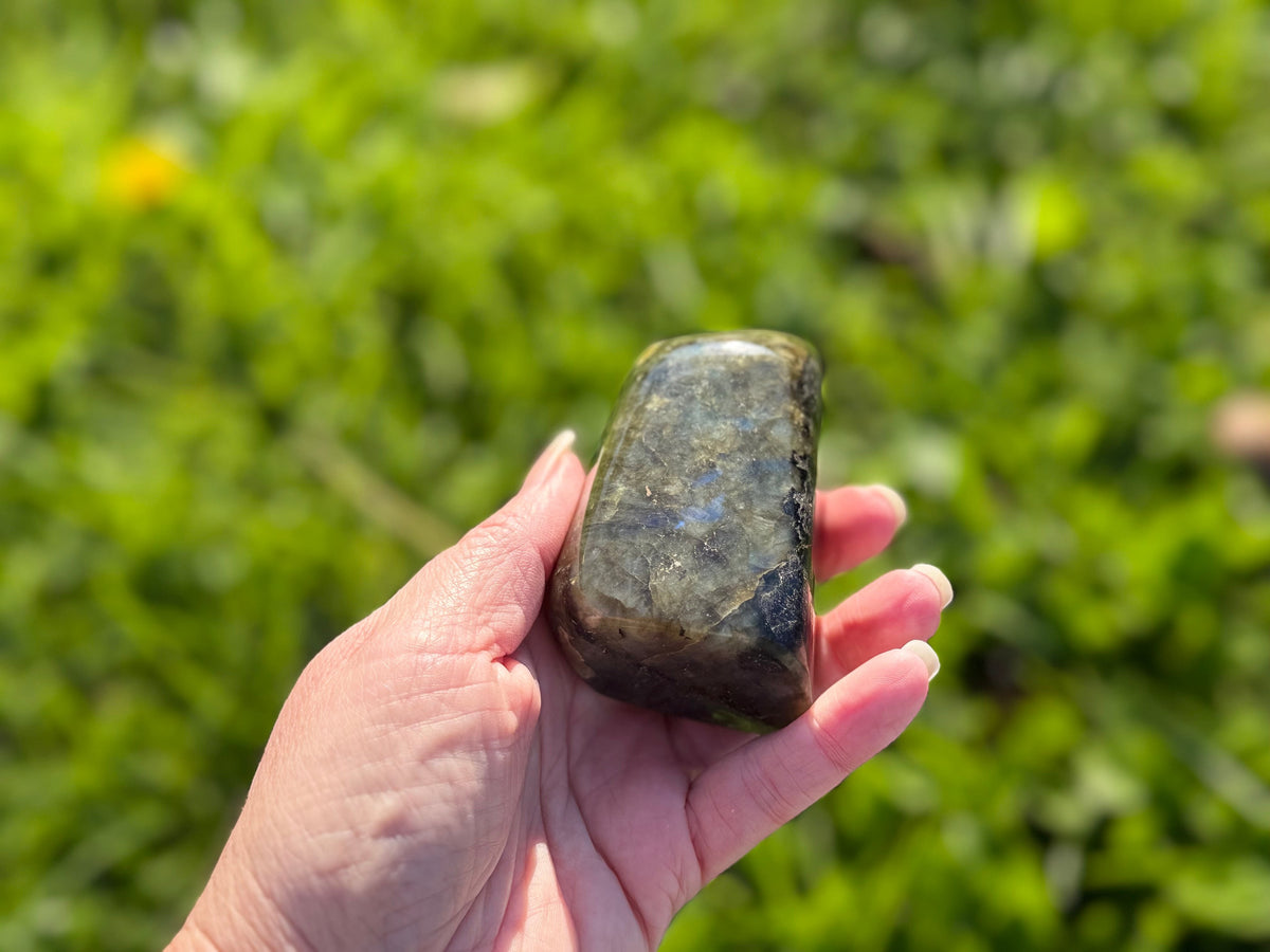 Treasured Energies hand-polished labradorite freeform crystal held in hand against a blurred green outdoor background.
