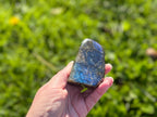 Treasured Energies polished labradorite freeform crystal held in hand, showing blue labradorescence against green grass.