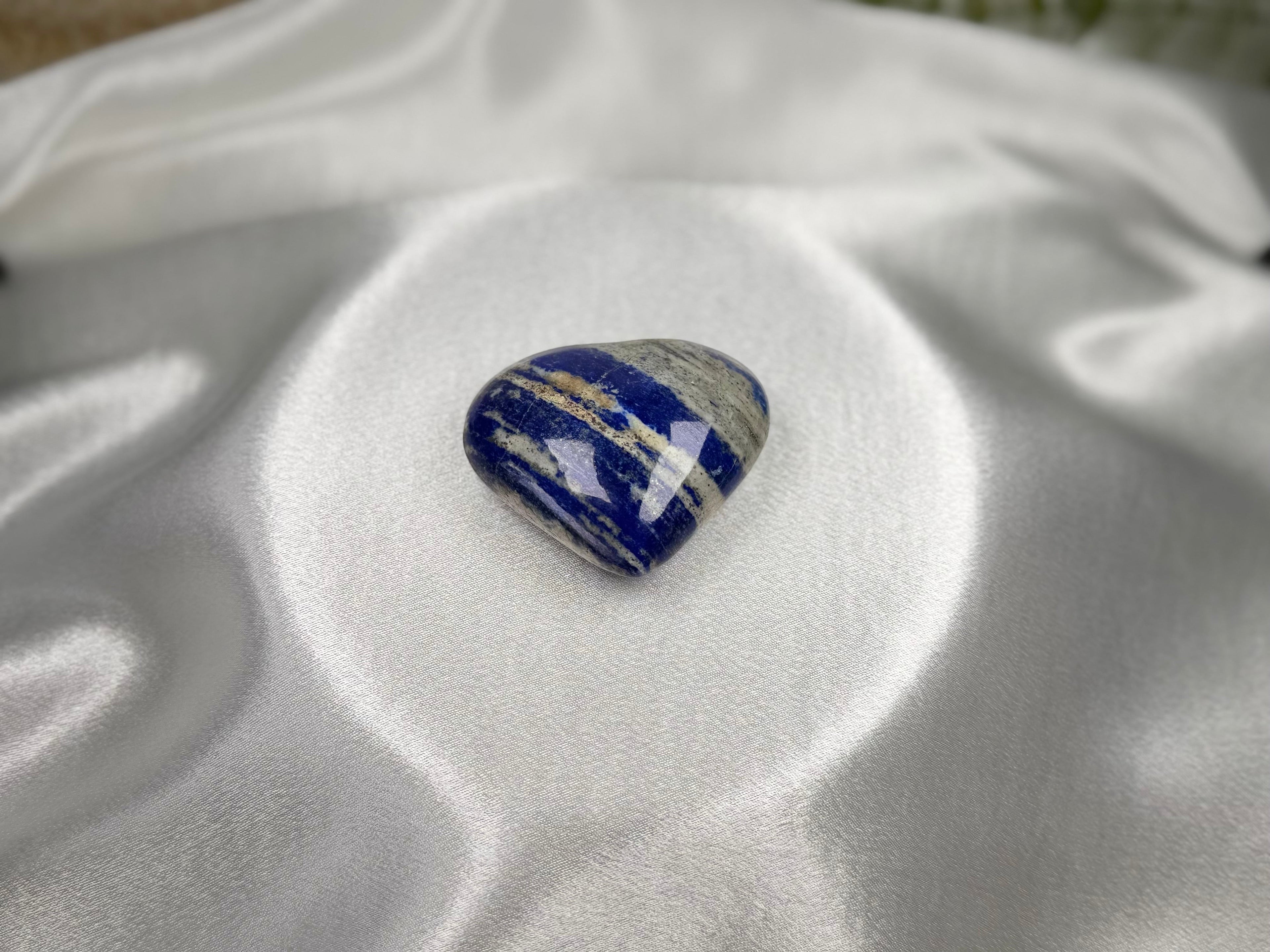 Treasured Energies polished Lapis Lazuli heart crystal with deep blue, white, and gold tones on white satin fabric.