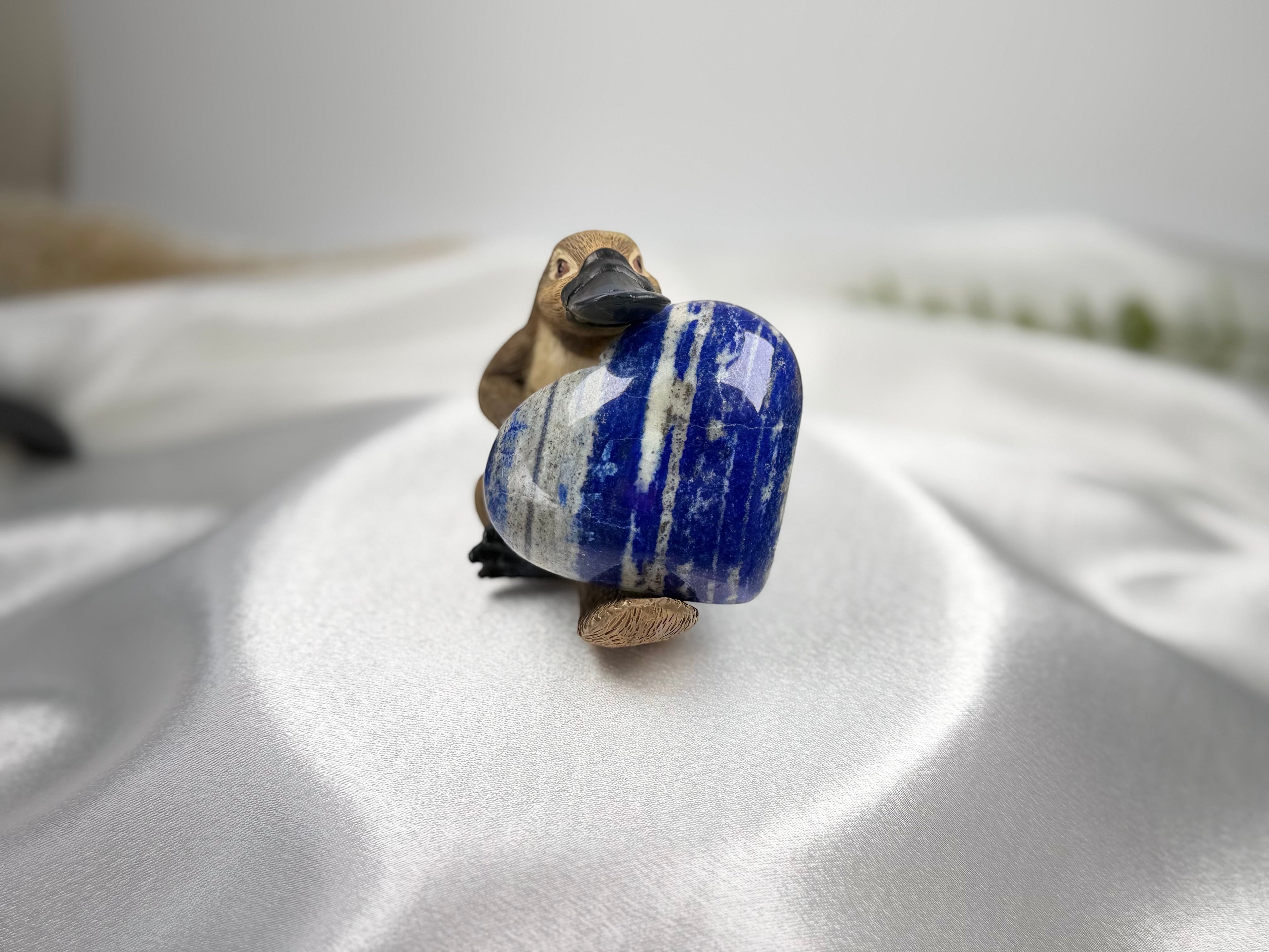 Treasured Energies polished Lapis Lazuli heart crystal held by a small wooden platypus figurine on white satin fabric.