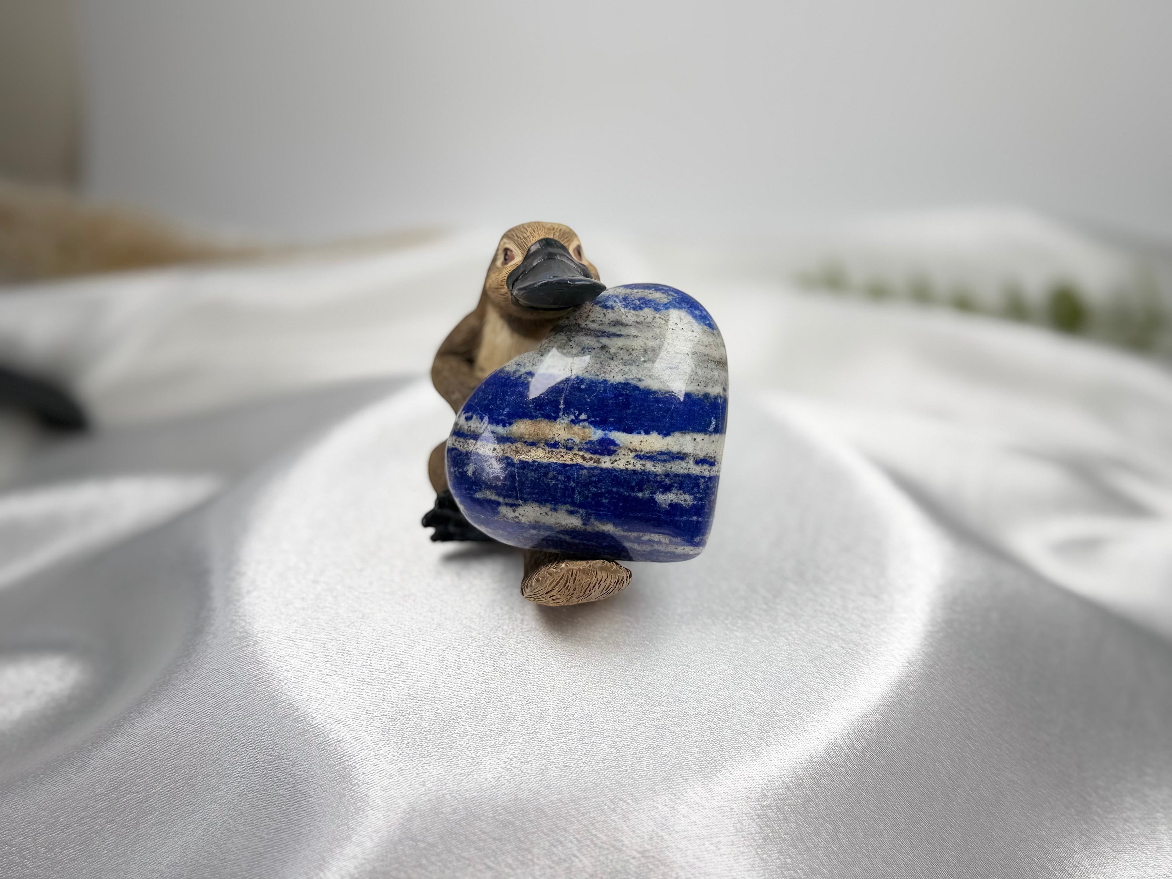 Treasured Energies polished Lapis Lazuli heart crystal with blue, white, and gold pyrite held by a small platypus figure.