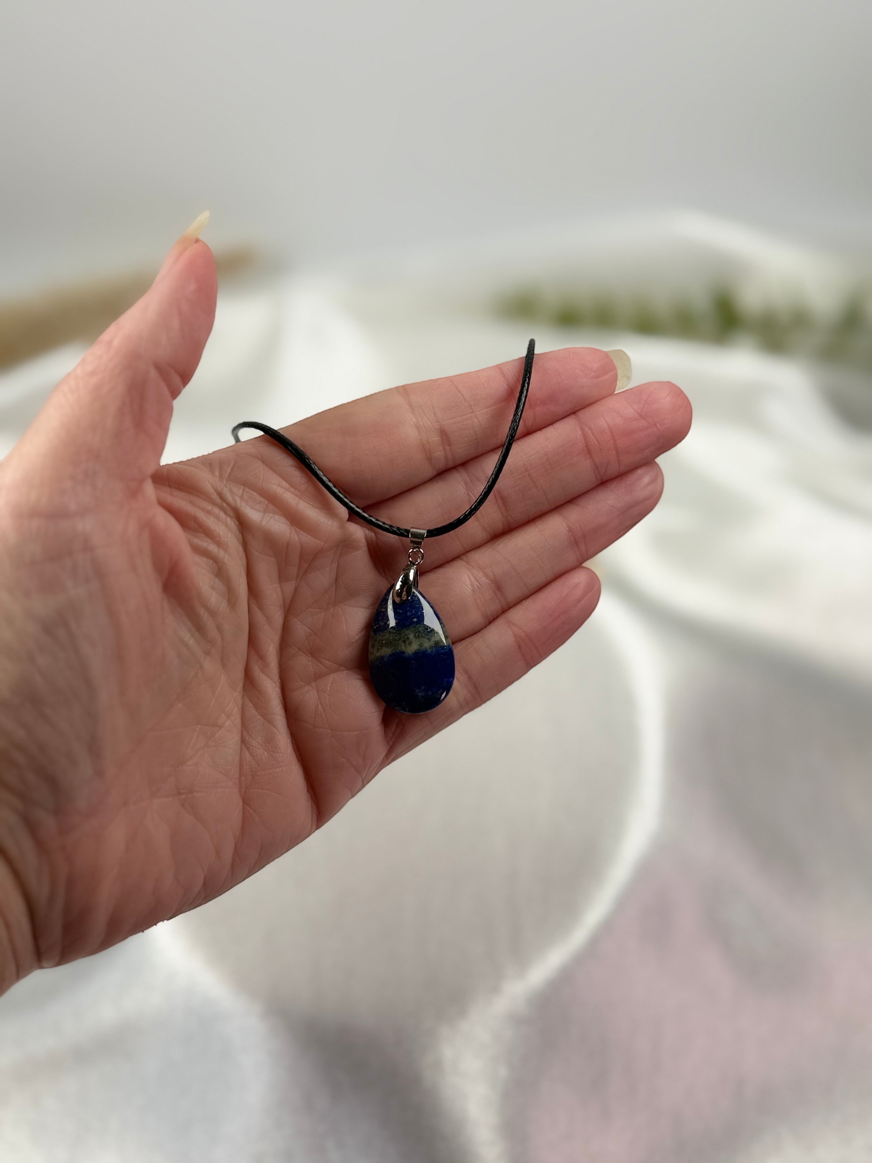 Treasured Energies Lapis Lazuli teardrop pendant necklace on a black cord held in a hand against a soft white background.
