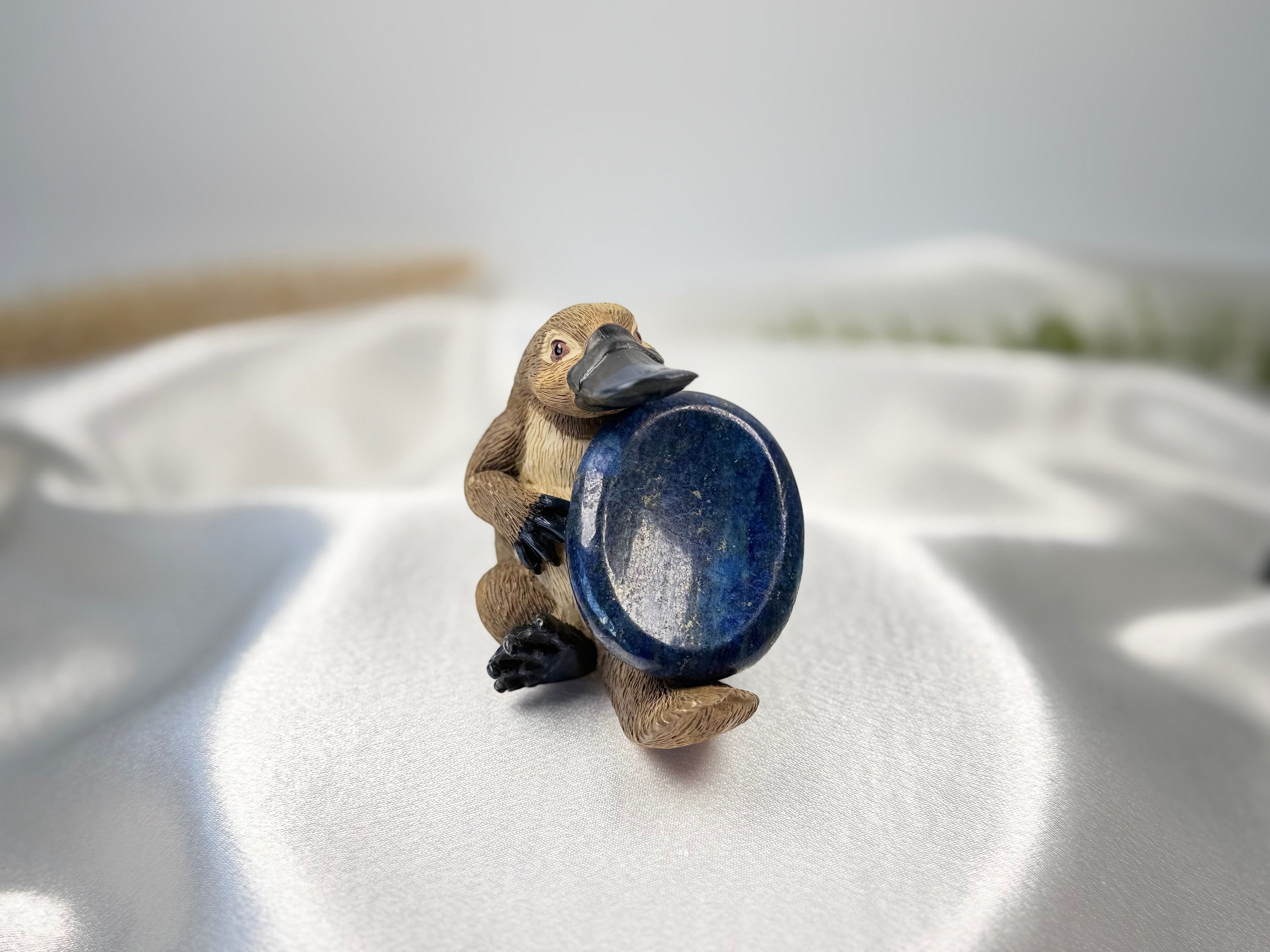 Treasured Energies Lapis Lazuli worry stone held by a small platypus figurine on a white satin fabric background.